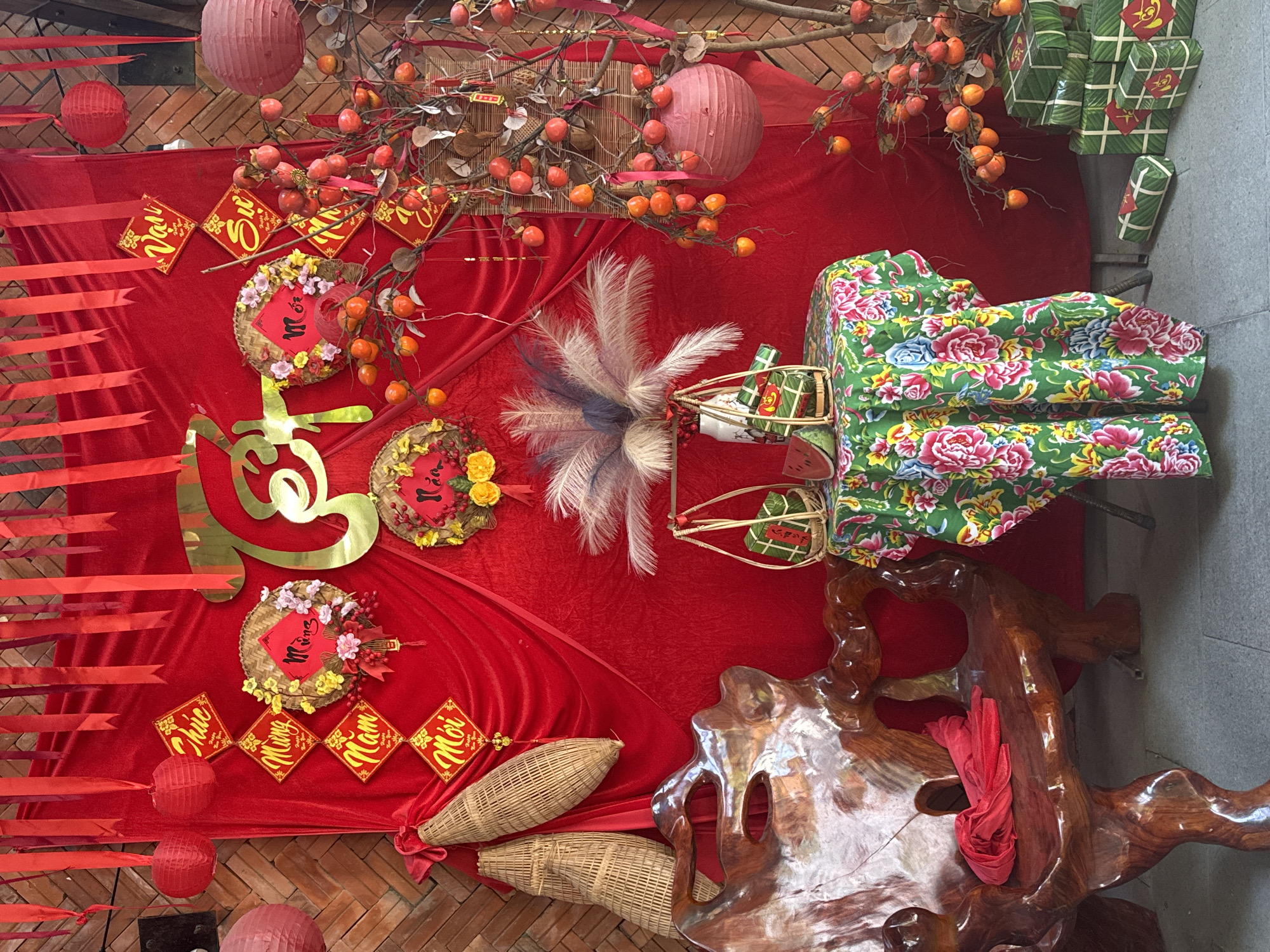 This is an image of a festive display, likely for a Vietnamese holiday such as Tết. The scene is dominated by vibrant red fabric, decorated with various traditional items. Several red lanterns hang from the ceiling and are scattered around. Branches laden with bright orange persimmons are prominently displayed, symbolizing good fortune.
Objects include woven baskets, some containing what appear to be banh chung (sticky rice cakes wrapped in banana leaves), which are also stacked in the background. Decorative items like golden calligraphy and circular ornaments adorned with flowers and Vietnamese text are placed on the red fabric. The text visible on the ornaments translates to "Happy New Year" or "Mừng Năm Mới." A large, intricately carved wooden stand is partially visible on the right side of the image, draped with a red cloth.
The setting appears to be an indoor or covered outdoor space, possibly a shop or a home decorated for a celebration. The overall atmosphere is celebratory and festive, evoking a sense of tradition and good wishes.