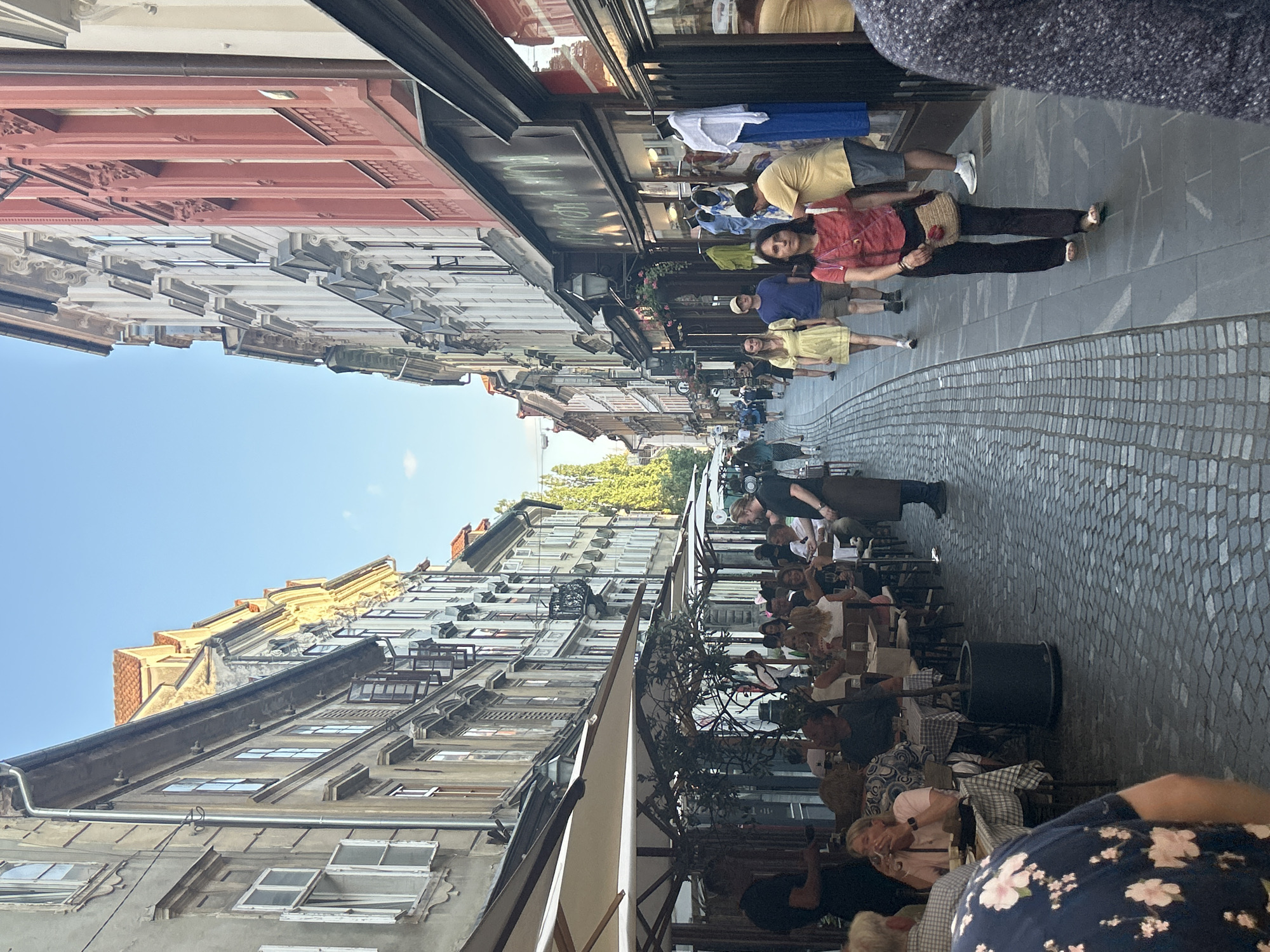 The image depicts a narrow cobblestone street lined with tall, old buildings with colorful facades. Outdoor seating areas for restaurants and cafes occupy a significant portion of the street. Several people are visible, some seated at tables and others walking along the street. The atmosphere appears to be that of a vibrant European city during the daytime, with clear skies. The buildings exhibit architectural styles common in older European cities, featuring multiple stories, arched windows, and decorative elements. The street is paved with cobblestones, and awnings extend from the buildings over the outdoor seating areas. The scene is alive with activity, suggesting a popular spot for dining and strolling.