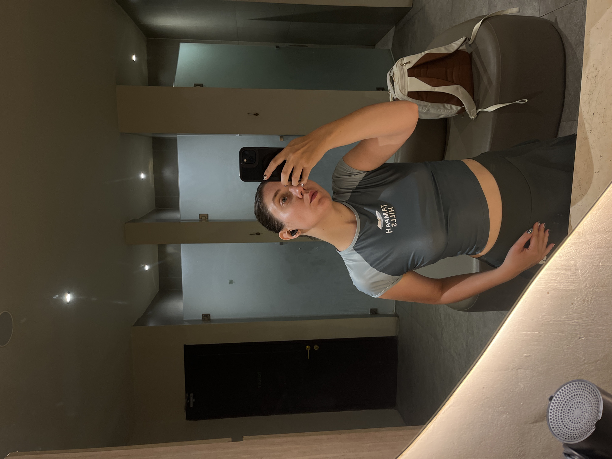 A woman is taking a selfie in what appears to be a locker room or changing area. She is holding a black smartphone up to her face and looking at the screen. She is wearing a gray and light blue athletic top and dark athletic shorts. In the background, there is a gray chair with a white backpack on it, as well as several locker doors and what looks like a shower area. The reflection suggests a mirror, and a hair dryer is visible. This is likely located in North Kuta, Indonesia.