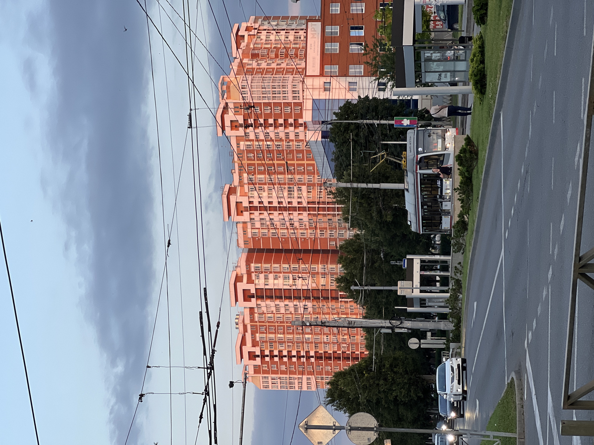 The image shows a street scene in Krasnodar, Russia. In the background, a large, modern residential building with a distinctive orange and white facade is visible, illuminated by the warm light of the setting sun. The building has many windows and balconies, and its top floors feature an unusual stepped architectural design. In the middle ground, a streetcar is stopped at a tram stop, with a few passengers visible inside and standing nearby. The street is paved and has multiple lanes, with cars driving on it. Overhead, a complex network of power lines crisscrosses the sky, suggesting an urban environment. Trees line the sides of the street, and a traffic signpost is visible on the left. The sky is partly cloudy, with a mix of blue and gray tones, indicating either early morning or late evening.