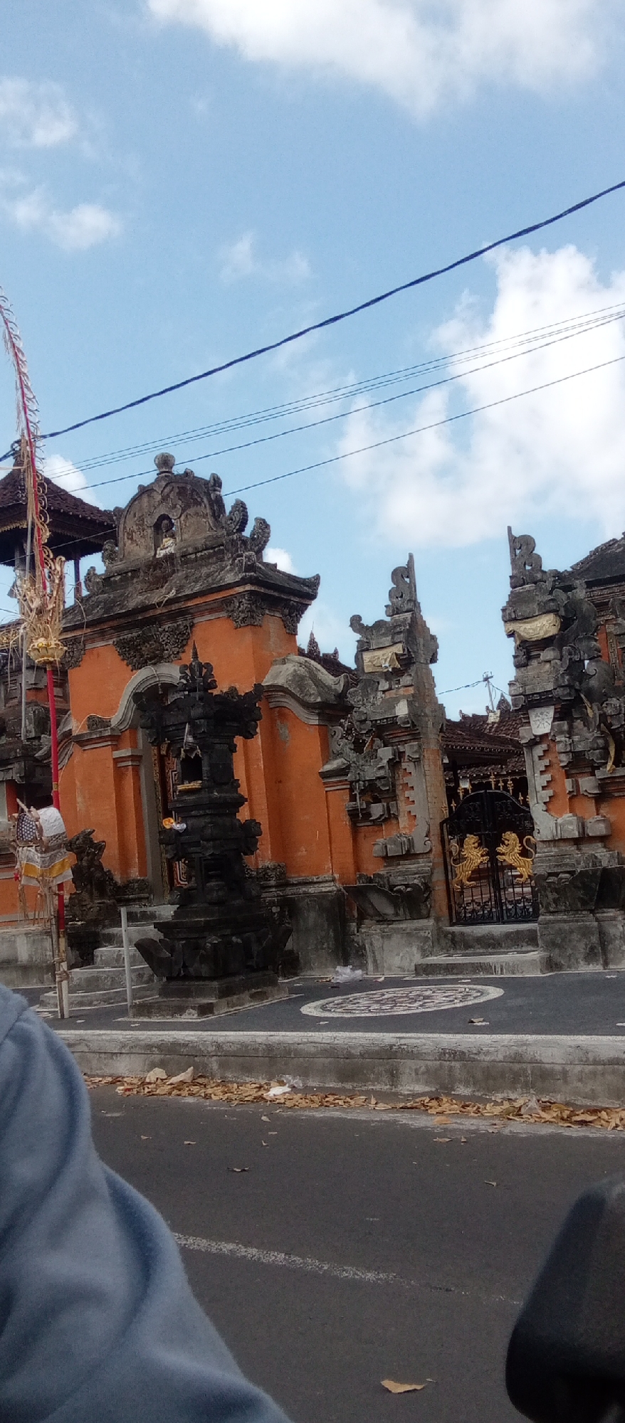 The image depicts the entrance to a traditional Balinese temple in Blahbatuh, Indonesia. The temple features ornate stonework with orange-colored walls and intricately carved grey stone accents. A prominent black stone sculpture stands in front of the main entrance, adorned with offerings. A large black metal gate with golden lion emblems is visible further back, leading into the temple grounds. Overhead, power lines crisscross against a bright blue sky with scattered white clouds. The foreground shows a paved road with scattered fallen leaves and a portion of a person's shoulder and arm, suggesting the photo was taken from a moving vehicle or while standing near the road.