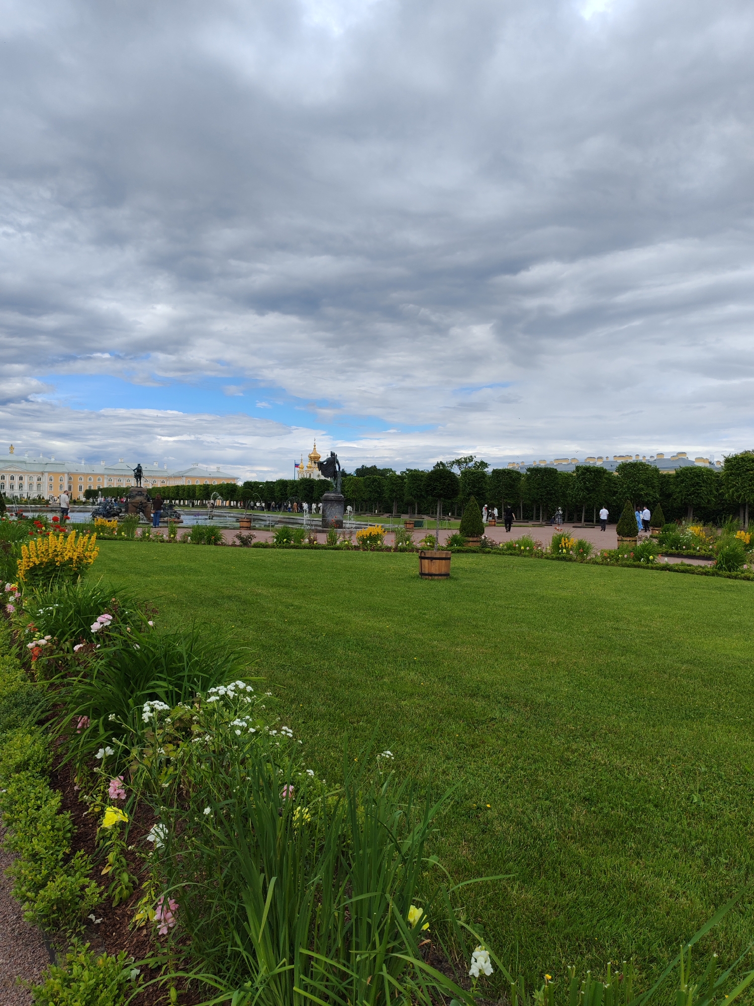 A wide shot captures a well-maintained garden and park area under a cloudy sky. In the foreground, a lush green lawn is bordered by a flower bed with a variety of colorful blooms, including yellow, pink, and white flowers, as well as green foliage. A gravel path runs alongside the flower bed. In the mid-ground, a circular fountain is visible, with water jets spraying upwards. Beyond the fountain, there is a row of manicured trees, and further in the distance, a grand, pale yellow building with a long facade is seen. Two golden-domed structures with spires rise from behind the trees and building. Several people are scattered throughout the park, walking and observing the scenery. A bronze statue on a pedestal is positioned near the fountain, and another statue of a figure with outstretched arms stands to the right of the fountain. A wooden barrel sits on the lawn in the mid-ground. The overall impression is one of an expansive and ornate public garden.
