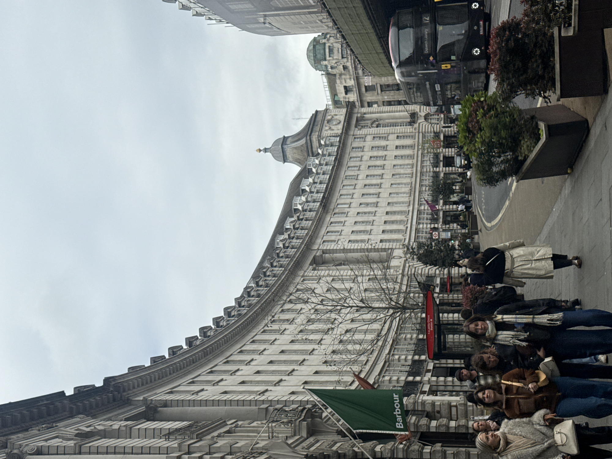 This photograph shows a street scene in London, United Kingdom, featuring the curved façade of a large, ornate building. A group of people, mostly young women, are standing and walking along the sidewalk on the right side of the frame. A large, black double-decker bus is parked near the curb. The building has rows of windows and intricate architectural details, with a dome visible on the upper levels. A green Barbour sign is attached to the building near the sidewalk. The sky is overcast.