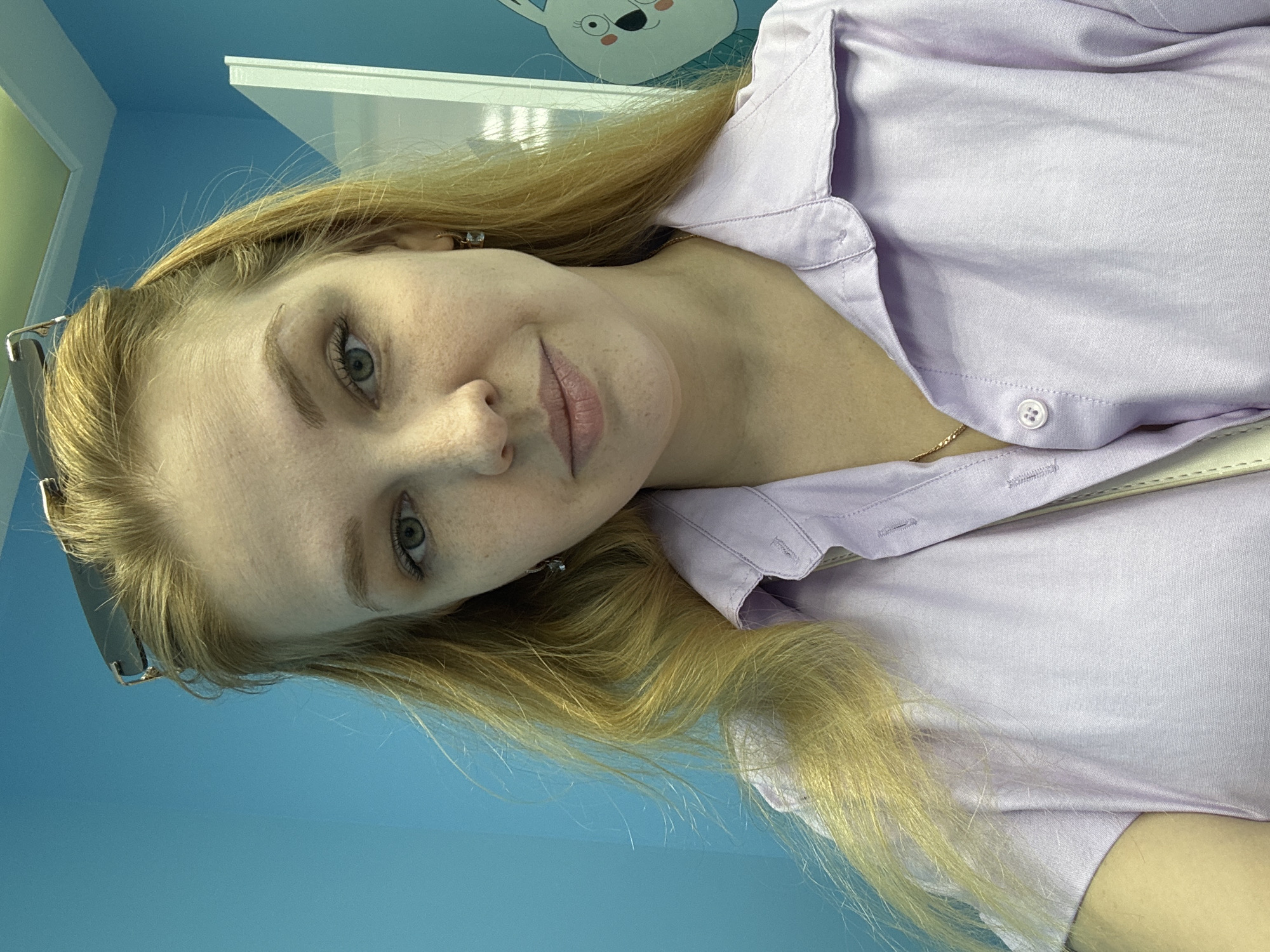 A young woman with blonde hair and fair skin, wearing a light purple collared shirt, poses for a selfie. She has freckles across her nose and cheeks and is wearing small hoop earrings. Sunglasses are perched on top of her head. The background consists of a bright blue wall with a white partition to the right, upon which a cartoon drawing of a rabbit is visible. The image appears to be taken indoors, possibly in a domestic or commercial setting. The location is not specified within the image itself.