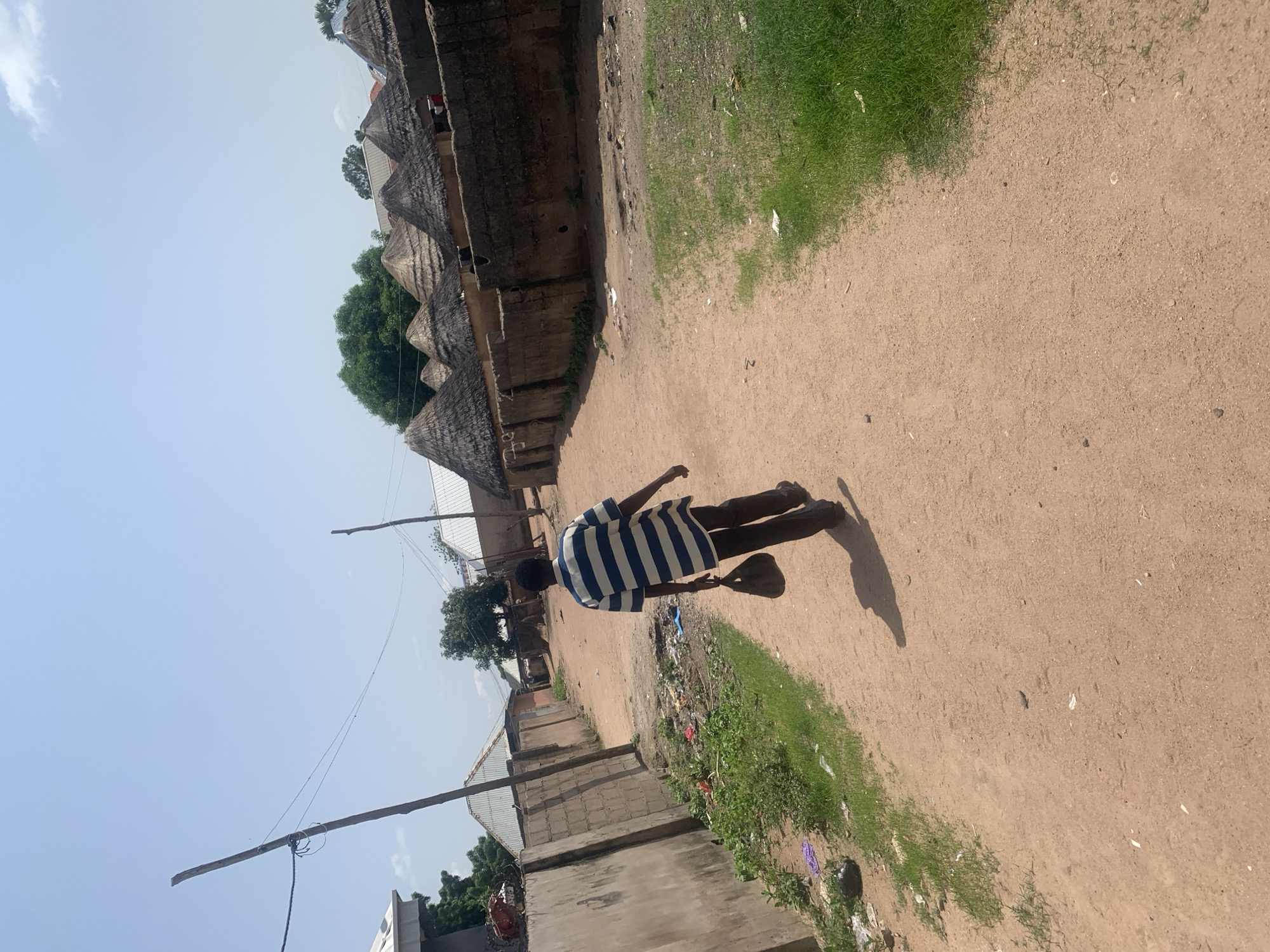 The image depicts a person walking away from the camera down a dirt path in a village setting, likely in Jalingo, Nigeria. The person is wearing a blue and white striped t-shirt and dark pants. They are casting a distinct shadow on the sandy ground. The path is lined on one side by traditional thatched-roof huts, characteristic of rural architecture in the region. Some of these huts appear to be made of mud bricks with conical straw roofs. On the other side of the path, there is a strip of green grass with some scattered litter. In the background, more buildings and trees are visible. The sky is a clear, pale blue, suggesting a bright, sunny day, likely late morning or early afternoon. There are power lines strung across the scene, supported by wooden poles, indicating some level of infrastructure. The overall impression is one of everyday life in a rural Nigerian community.