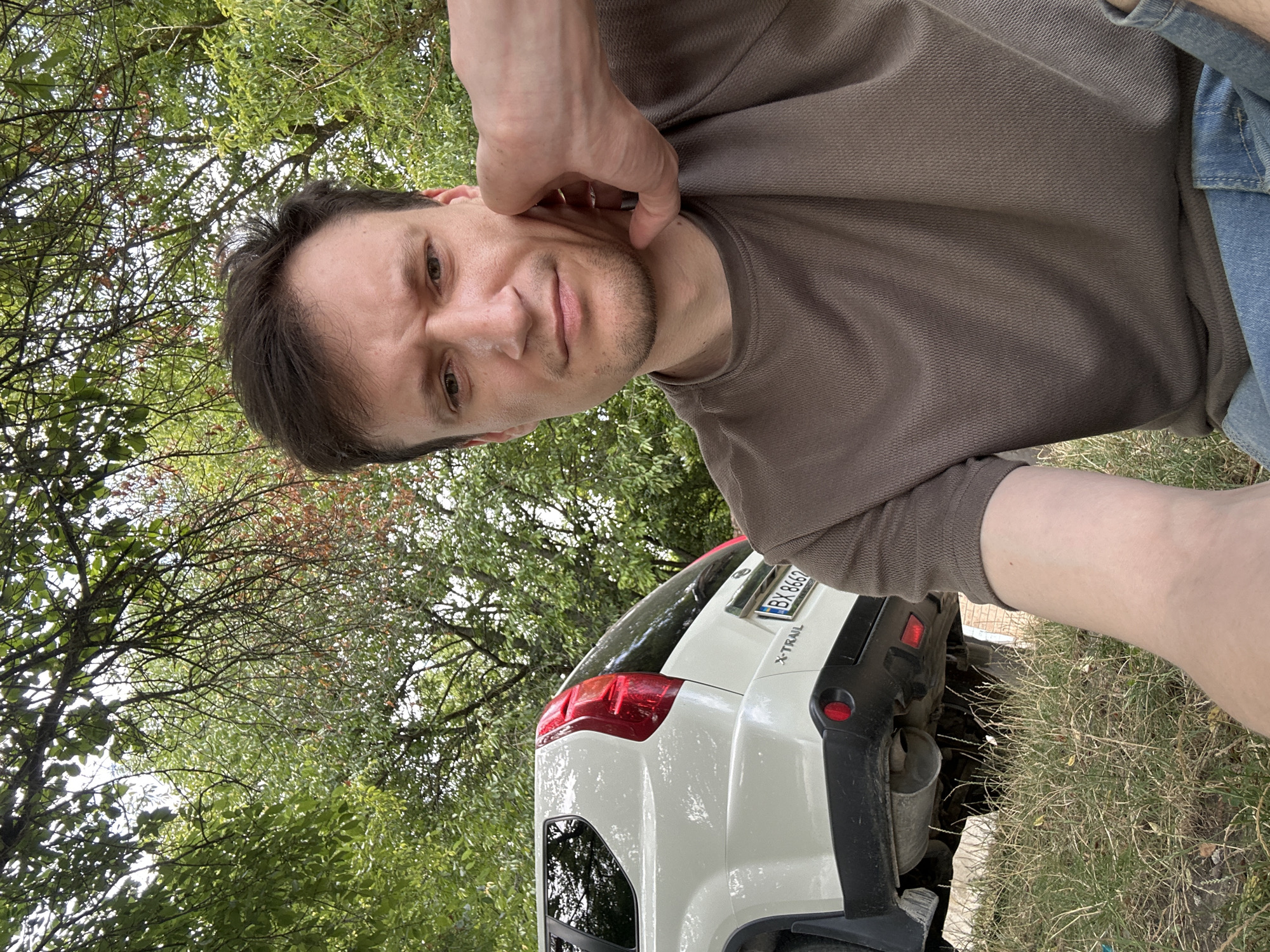 A man with dark hair and brown eyes is taking a selfie outdoors. He is wearing a brown t-shirt and blue jeans. His right hand is touching his chin. In the background, a white Nissan X-Trail SUV is parked on a grassy incline. The vehicle's license plate reads "BX 8662". The scene is set amidst trees with green and reddish foliage, suggesting an outdoor, possibly park-like environment, located in or around the city of Makiv, Ukraine.