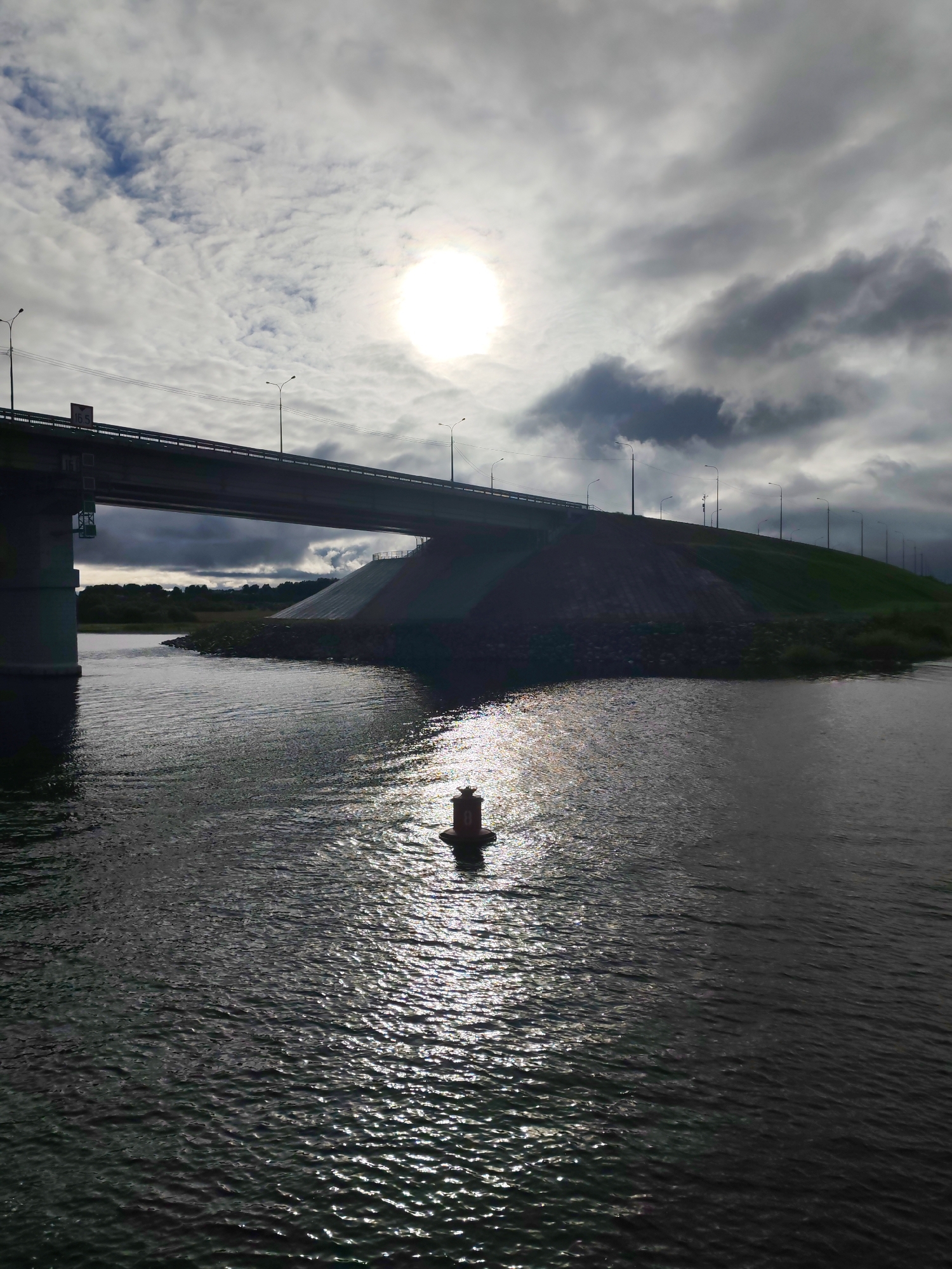 A view of a bridge in Нифантово, Russia. The image is taken from the water, showing a waterway in the foreground with a single red buoy floating on the surface. Sunlight reflects off the water. A bridge spans the waterway, supported by concrete pillars and flanked by a green embankment. Streetlights line the top of the embankment. The sky is overcast with the sun shining through the clouds.