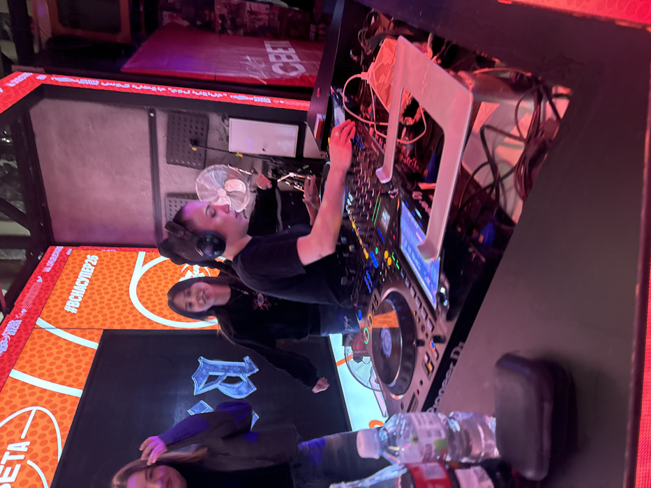The image displays an indoor scene at Sveta Bar, located at Ulitsa Marata, 2, Moscow, Russia. A female DJ, estimated to be in her mid-20s to early 30s, with dark hair tied back and multiple tattoos on her left arm, is actively engaged at a DJ booth. She wears large black over-ear headphones and a black short-sleeved top, with her right hand manipulating controls on a Pioneer DJ mixer featuring colorful illuminated buttons and a blue display screen. A small white desk fan is positioned behind her, directed forward.
To the immediate left of the DJ, a second female of similar age, with dark hair and wearing a dark long-sleeved top with a light-colored eye graphic, stands and observes. Further left, partially visible, is a third female, also in her mid-20s to early 30s, with light brown hair, dressed in a dark jacket over a dark top and light blue jeans, with her left arm raised.
On the black surface of the DJ booth, to the left of the equipment, are two plastic water bottles (one with a blue cap) and a bottle of Coca-Cola. A small, rectangular black case rests nearby. To the right of the DJ equipment, a silver-colored metal laptop stand is present, with various cables visible underneath. A folded red face mask is also on the booth surface near the laptop stand.
The background features a large LED screen displaying an orange, textured pattern resembling a basketball. White Cyrillic text "СВЕТА" is visible on the upper left, and "#ВСЯСУПЕР26" is on the upper right. Below "СВЕТА," a stylized blue letter, resembling a Cyrillic "В" or English "B," is prominent against a dark background. To the right of the main screen, a narrower vertical LED panel displays "СВЕТА" in white Cyrillic text on a red background. The entire DJ setup and screens are integrated into a dark, industrial-style metal framework. The environment is dimly lit by ambient light, contrasting with the bright, vibrant illumination from the LED screens and DJ equipment.