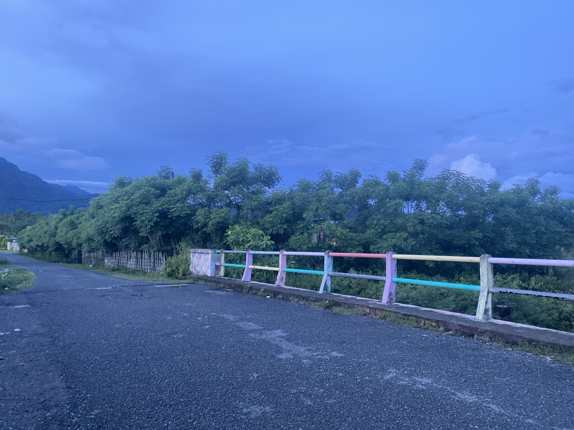 The image depicts a twilight scene on a road in Aneuk Paya, Indonesia, with a mountain range visible in the distance. A dark asphalt road curves to the left, leading into a densely vegetated area. On the right side of the road, a concrete railing separates the road from the vegetation. The railing is decorated with brightly colored horizontal bars in pastel shades of pink, yellow, green, and purple, interspersed with concrete posts. The sky above is a deep, moody blue, with hints of clouds. The overall impression is one of a rural or semi-rural setting as dusk settles.