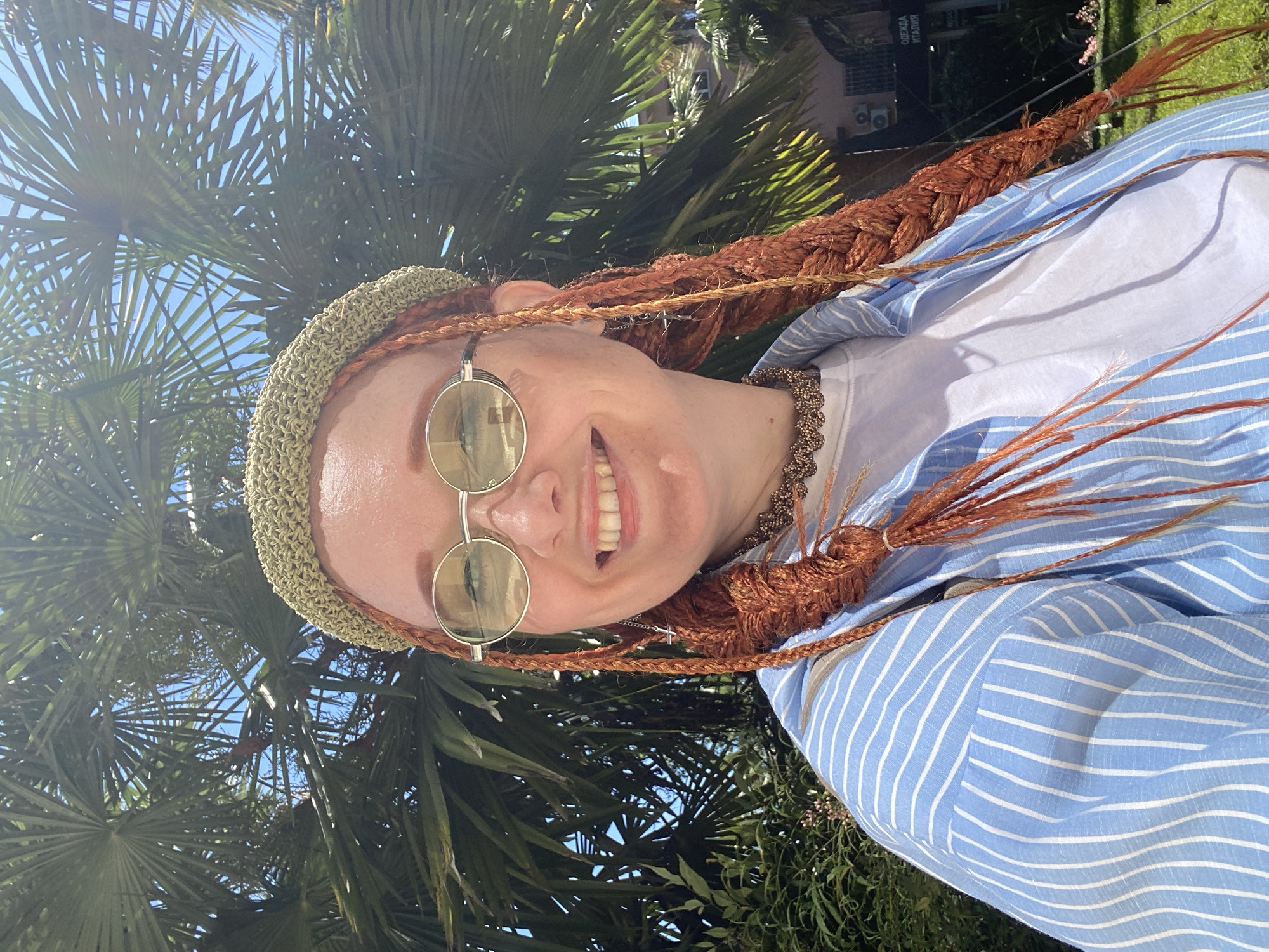 A close-up selfie shows a woman with auburn braids and a green knitted hat, wearing round, gold-rimmed sunglasses and a blue and white striped button-down shirt over a white t-shirt. A decorative choker necklace is visible. The background features large palm fronds and a bright blue sky. A building with Cyrillic signage indicating "ОДЕЖДА ИТАЛИЯ" (Clothing Italy) is visible in the distance, along with some greenery and sunlight. The overall impression is an outdoor setting, likely in a warm climate, with elements suggesting a tourist location.