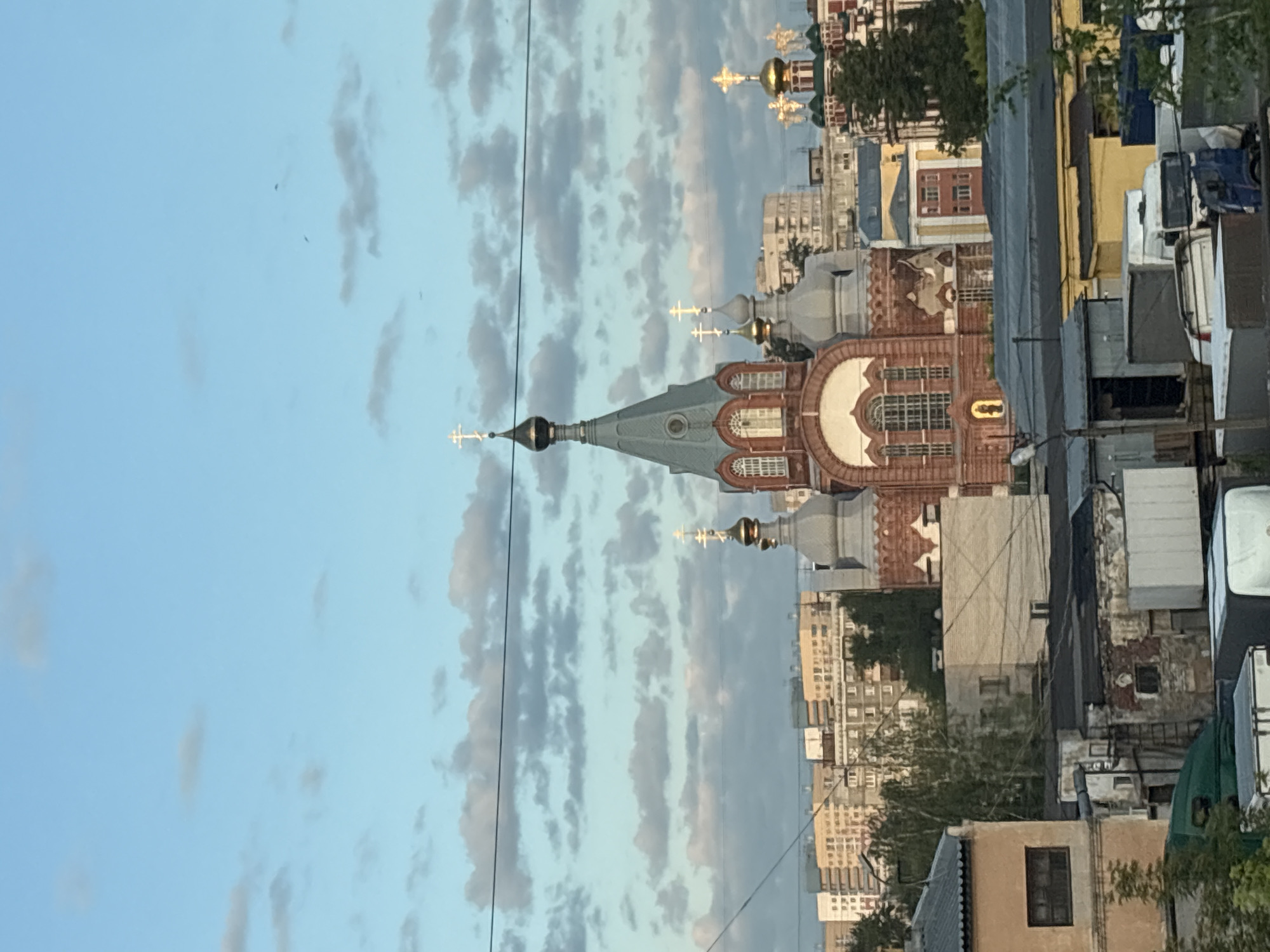 The image captures a view of the Orthodox Church of the Resurrection of Christ in Nizhny Novgorod, Russia. The church, with its distinct red brick facade and multiple domes topped with golden crosses, is the central focus. It is surrounded by a mix of urban elements, including multi-story residential buildings, smaller industrial or storage structures, and some greenery. The sky above is a soft blue, dotted with wispy, light grey clouds, suggesting it might be late afternoon or early evening. There are no people or animals visible in the frame, and the scene is static. The overall impression is one of a cityscape with a prominent religious structure.