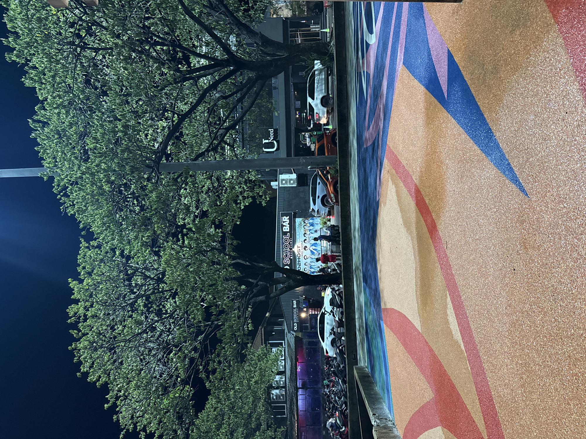 This is a night-time, eye-level, medium shot of a street scene in the Koh Samui District of Thailand. In the foreground, there is a colorful, textured surface with abstract designs in shades of orange, red, blue, and pink, resembling painted asphalt or a similar outdoor flooring material. Beyond this, a large number of parked motorcycles are visible, lining the sidewalk. Behind the motorcycles, there are several buildings. The building on the left has a sign that reads "OLD SCHOOL BAR". Further to the right, a building has a large sign with the logo "U Need" and text that appears to be in English. To the right of the "U Need" building, there is another sign that says "OLD SCHOOL BAR" and "2HOST CLUB". A group of people, appearing to be young men in blue outfits, are posing for a photo in front of a poster or display. There are several cars on the road in front of the buildings, including a white sedan and a white SUV. Two people are standing near the poster. The scene is illuminated by artificial lighting, with large trees with green foliage casting shadows. The sky is dark, indicating it is nighttime.
