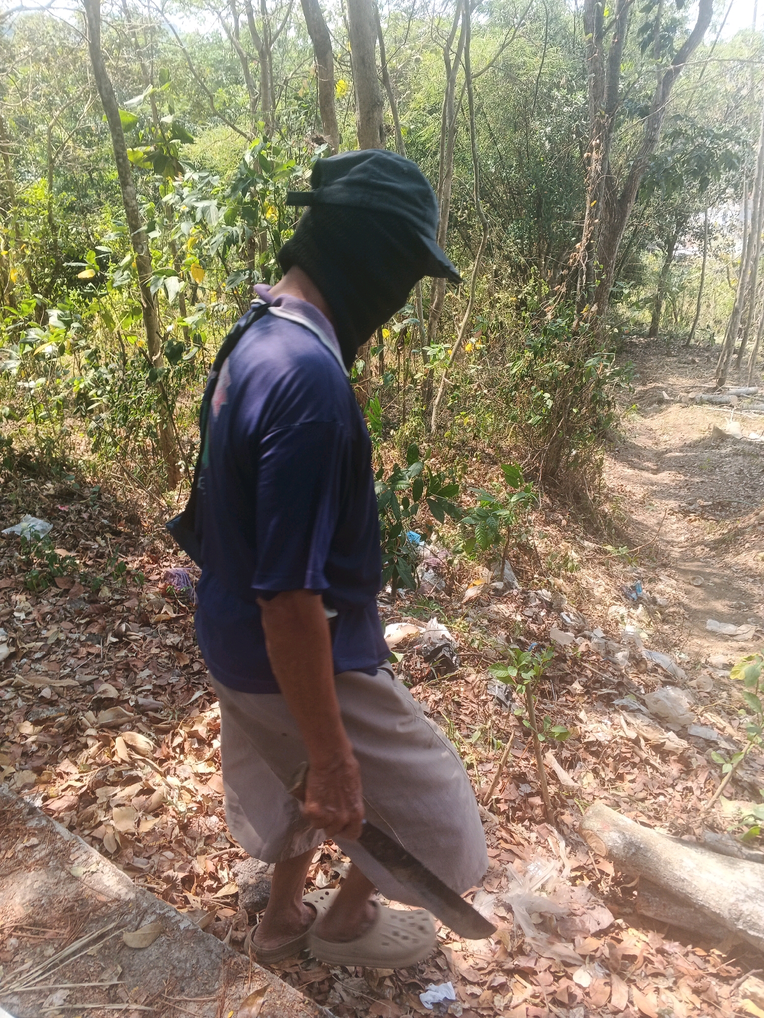 A person wearing a dark blue t-shirt, light brown shorts, and a dark hat that covers their face is walking on a dirt path in a wooded area. The person is holding a machete in their right hand. The ground is covered with dry leaves, and there is some litter scattered around. The trees are tall and thin, and the sunlight is filtering through the leaves, creating shadows on the ground. The scene appears to be in a rural or undeveloped area, possibly on the outskirts of Bandarjo, Indonesia.