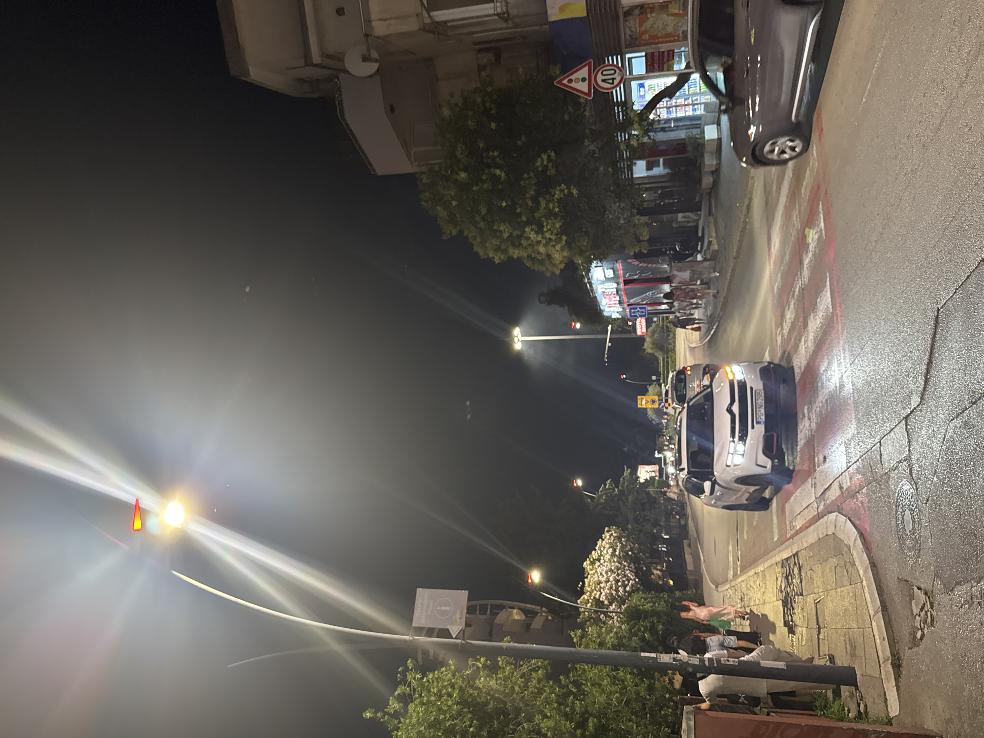 This is a nighttime street scene in Budva, Montenegro. A white SUV is stopped at a pedestrian crossing marked with red and white stripes. To the right, a gray SUV is partially visible, also near the crossing. In the background, there are buildings with lights on, including what appears to be a restaurant with neon signage. Trees line the street, and streetlights illuminate the scene. Several people are standing on the sidewalk to the left, waiting or walking. One person is holding a green bag. A "Tourist Information Point" sign is mounted on a lamppost. A "40" speed limit sign is visible on the right side of the street. The road surface appears wet, suggesting recent rain.