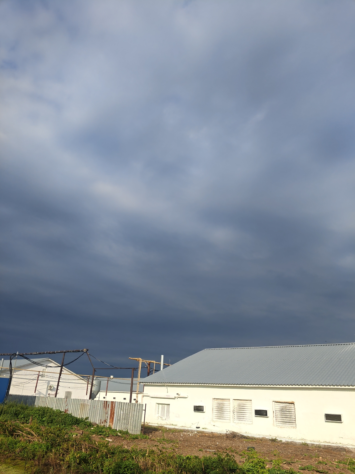 The image shows a modern white building with a corrugated metal roof, situated in an industrial area. The building has several windows with louvers and some smaller vents. A corrugated metal fence separates the building from a grassy area in the foreground, which is overgrown with weeds and dry vegetation. In the background, there are other industrial structures, including what appears to be a storage tank and various pipes and electrical conduits. The sky is overcast with dark, heavy clouds, suggesting an impending storm. The location is identified as Platoschino, Russia. There are no people or animals visible in the image.
