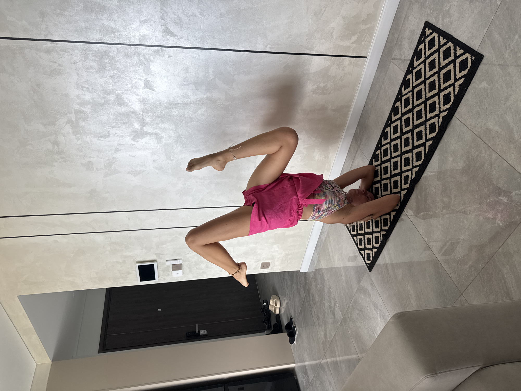 A woman in a pink skirt and patterned top is lying on a black and beige patterned rug in a room. Her legs are raised and bent at the knees. The room has gray tiled floors and light gray textured walls with dark linear accents. A black door and various shoes are visible near the wall. Đà Nẵng, Vietnam is not identifiable in the image.