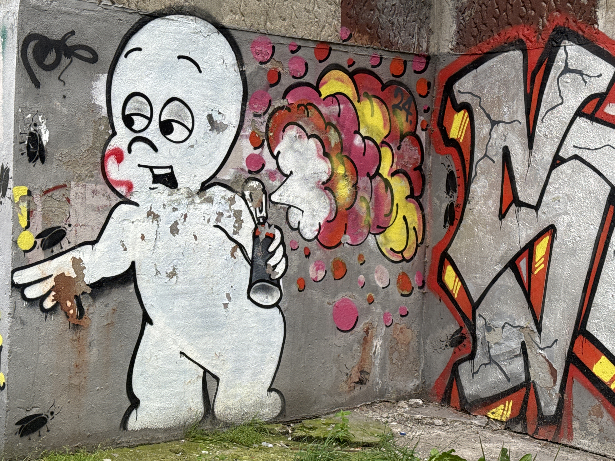 The image captures a section of graffiti art on a weathered concrete wall in Kaliningrad, Russia. The artwork features a cartoonish, white ghost-like figure on the left, holding what appears to be a spray can and emitting colorful smoke. The smoke is depicted in shades of red, pink, and yellow, with circular accents in pink and orange. To the right of the ghost, large, stylized block letters, possibly initials, are spray-painted in silver with red and yellow highlights, outlined in black. Several small, black insect-like figures are visible scattered across the lower portion of the wall, near the ground. The wall itself shows signs of age and damage, with peeling paint and rust stains. The ground in front of the wall is covered with sparse green grass and some debris. The scene suggests an urban environment, likely an outdoor public space where graffiti is common.