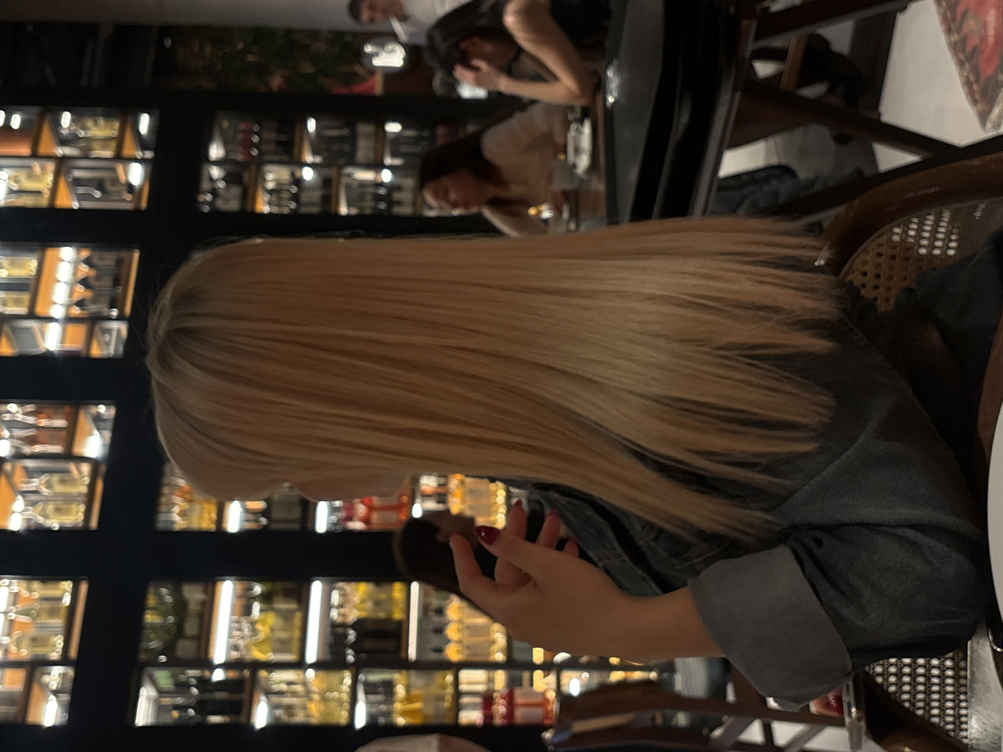 A woman with long, blonde hair sits at a table in what appears to be a restaurant or bar, possibly in Yerevan, Armenia. Shelves behind her showcase numerous bottles, suggesting a wide selection of alcoholic beverages. She is holding a dark-colored object, potentially a phone, in her hands, and her fingers have red nail polish. The woman is wearing a dark denim jacket or shirt. Other individuals are visible in the background, seated at nearby tables. The image is taken at a close-up perspective, focusing on the woman and the immediate surroundings.