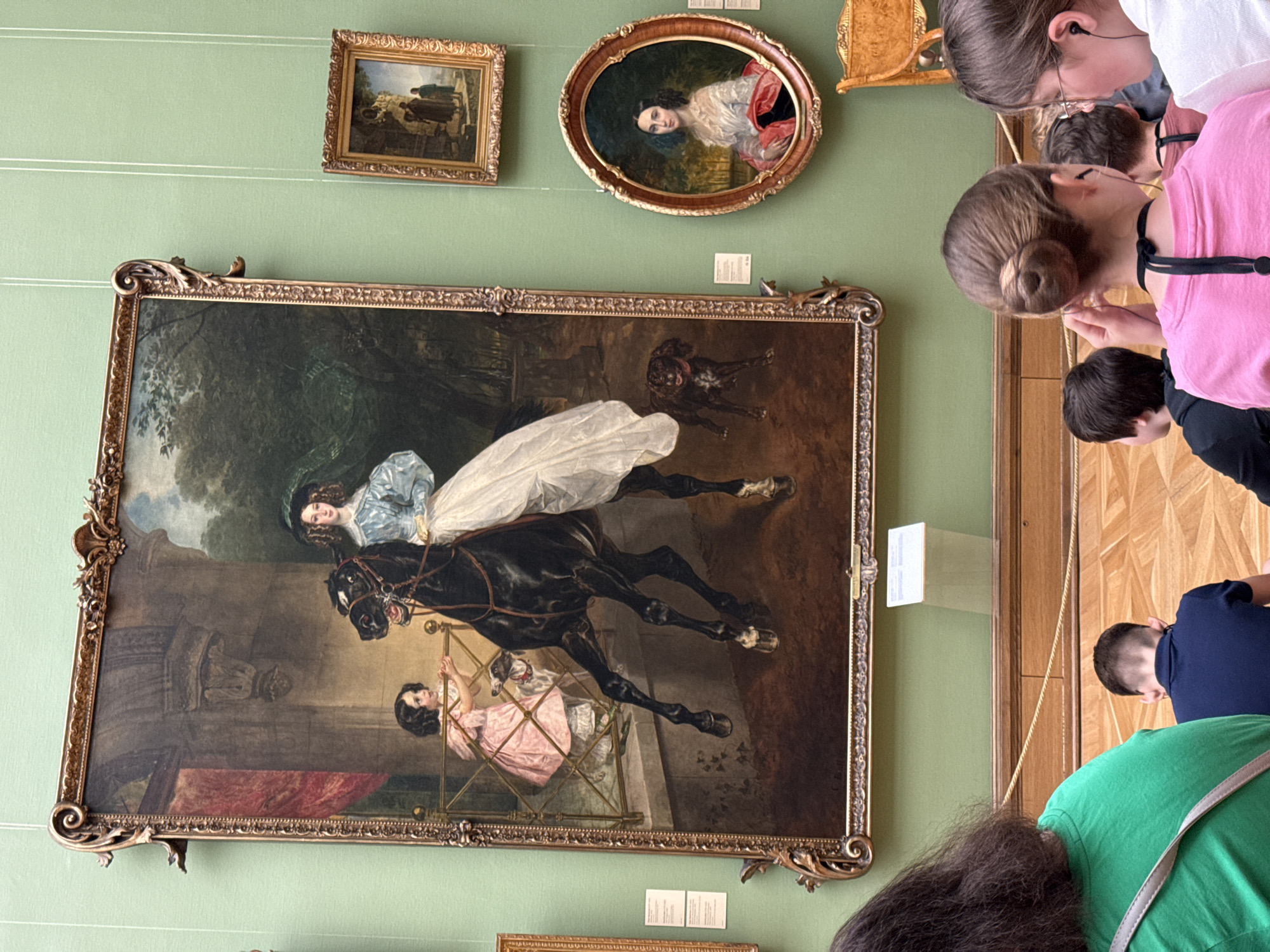 The image shows a large, ornately framed painting on a pale green wall, surrounded by other smaller framed artworks. The painting depicts two women and a dog. One woman in a blue dress with a white skirt is riding a black horse. A young girl in a pink dress sits in what looks like a cage in the lower right corner. A dark-colored dog is positioned behind the horse. Several people are standing in front of the painting, likely viewing it. The location is likely a museum or art gallery in Moscow, Russia, given the nature of the artworks and the presence of viewers.