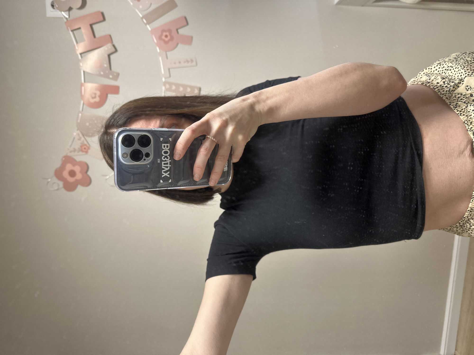 A woman is taking a mirror selfie. She is holding a smartphone in her right hand. The phone case is translucent and displays text in Cyrillic script along the side. She is wearing a black short-sleeved shirt and patterned pants. On her right ring finger, she has a thin gold ring. Behind the woman, a garland with pink decorative letters and shapes is visible on the wall. The location is Votkinsk, Russia.