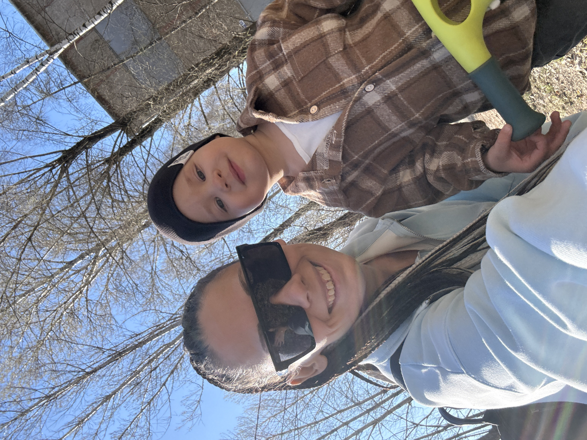 This image captures a cheerful outdoor moment in Rusanovo, Russia, featuring an adult and a child. The photo is a selfie taken from a low angle, looking upwards towards a bright, clear blue sky and bare tree branches.
In the foreground, an adult, likely female, is smiling broadly. She wears large black reflective sunglasses and a light blue hooded top, and has dark hair styled in braids. Slightly behind and above her is a young child, appearing to be male, also smiling. The child wears a black beanie adorned with a white star emblem and a brown and white plaid shirt over a white t-shirt. He is holding a bright yellow and dark green toy, resembling a small shovel or scoop.
The background is dominated by the intricate network of bare tree branches against the clear blue sky, suggesting either late winter or early spring before the foliage has grown. The white bark visible on some trunks indicates the presence of birch trees. In the upper background, a dark grey or brown structure, possibly a roof or part of a building, is visible. The bright sunlight and the smiles of both individuals convey a positive and happy atmosphere. No explicit text is clearly visible, though the child's cap has a star emblem.