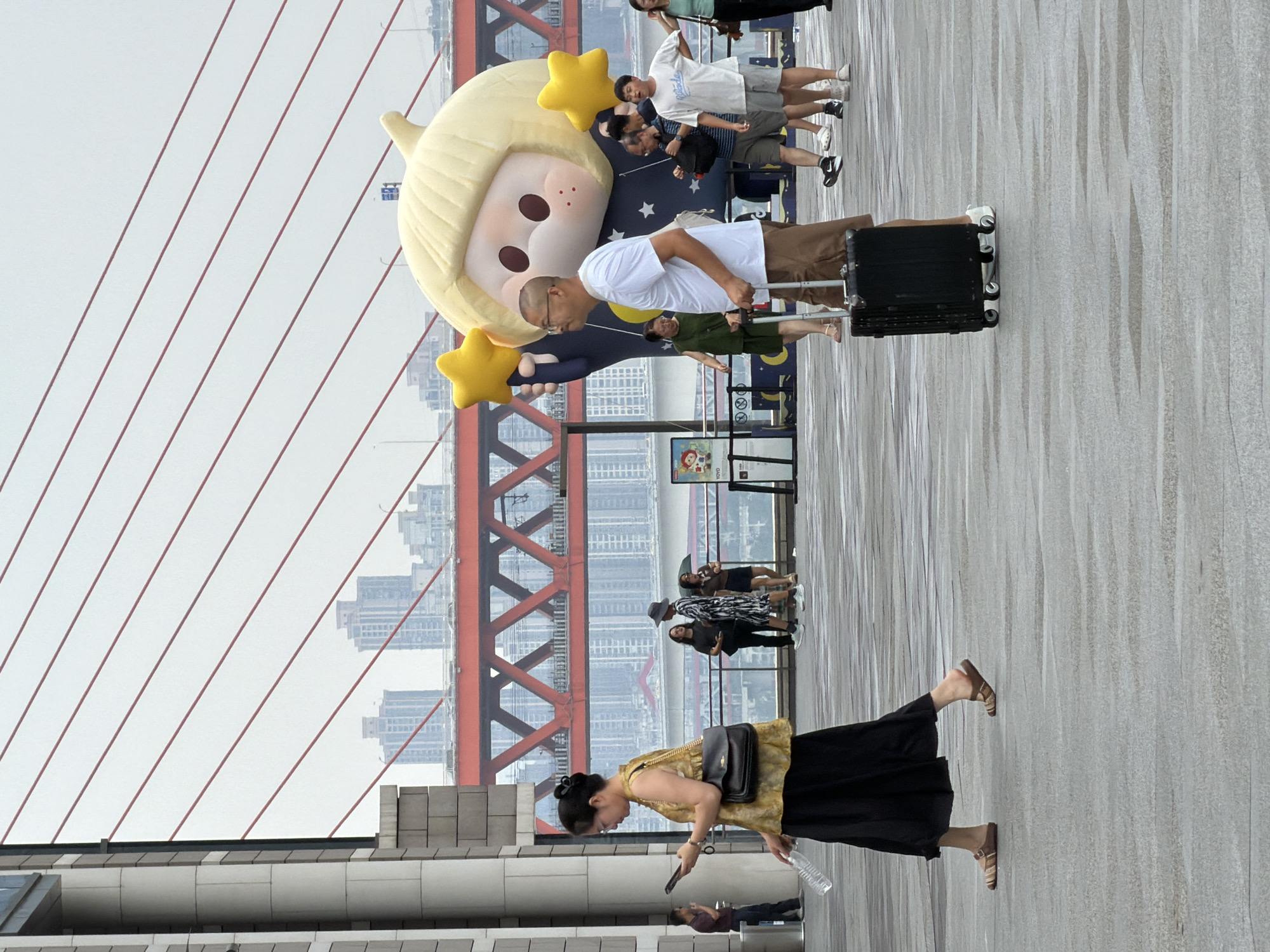The image depicts an outdoor scene on what appears to be a pedestrian walkway or bridge, possibly in the Yuzhong District of China, given the urban architecture and bridge structure visible in the background. The ground is wet, suggesting it has recently rained or is a damp day.
In the foreground, a woman in a yellow sleeveless top and a black skirt is walking towards the left, holding a clear plastic bottle in her right hand and looking at her phone. She is wearing brown sandals. To her right, a man in a white t-shirt and brown shorts is walking towards the right, pulling a black rolling suitcase. He is wearing glasses and looking ahead.
Behind them, a large inflatable cartoon character with blonde hair and star motifs is visible, holding yellow stars. Several other people are scattered throughout the background. Some are walking, and others appear to be standing and observing.
In the middle ground, a red bridge structure with intricate crossbeams dominates the background, spanning across a body of water or cityscape. The skyline behind the bridge features multiple tall buildings, indicating an urban environment. The sky is overcast.