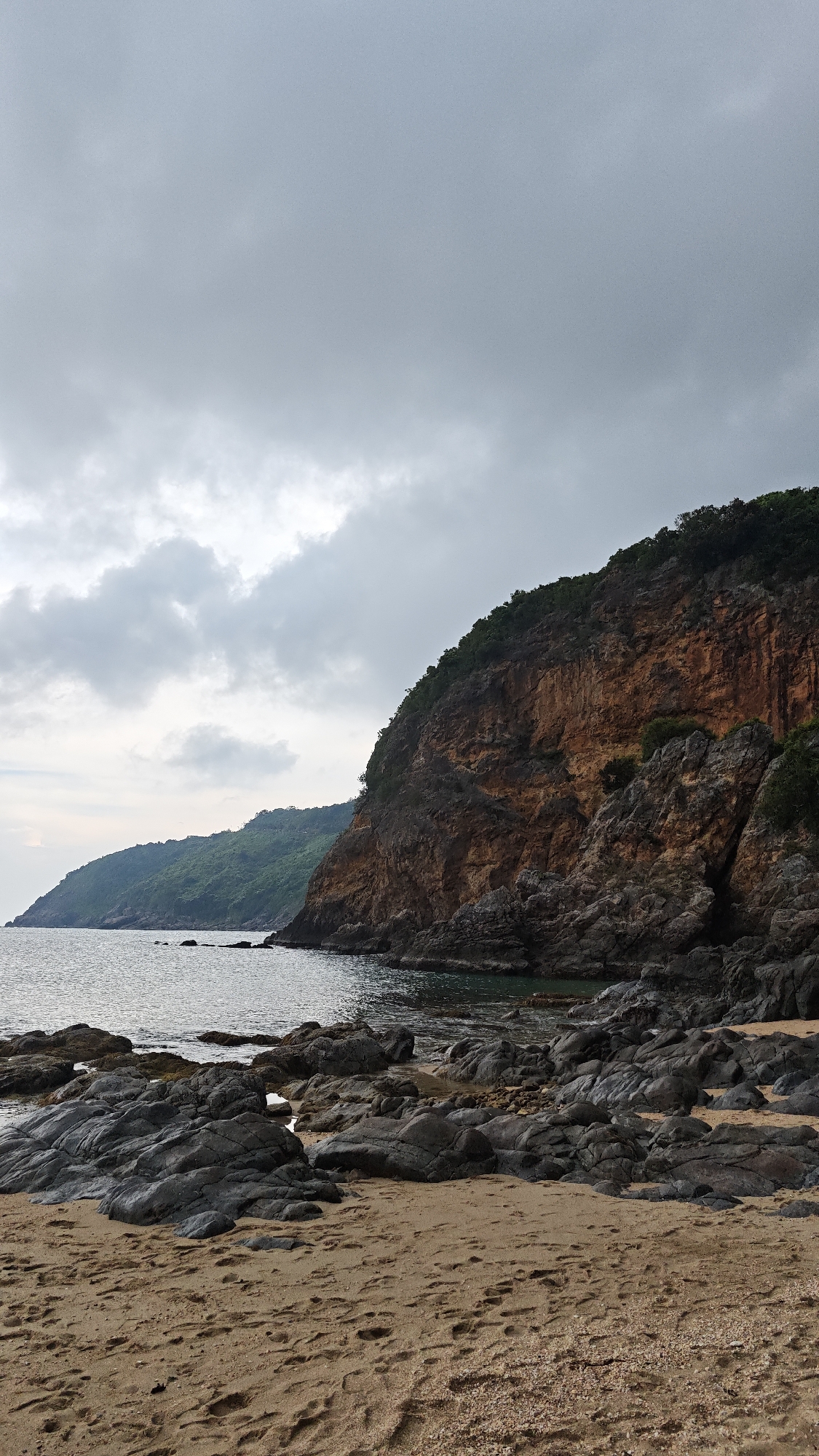 The image depicts a rocky beach in Đà Nẵng, Vietnam, under a cloudy sky. The foreground shows sand, merging with scattered dark grey rocks of varying sizes. In the middle ground, a cliff rises from the water's edge, its face a mix of dark grey rock and reddish-brown soil. Green vegetation covers the top of the cliff. The sea is calm. In the background, a similar but smaller land formation is visible, also covered with green vegetation. The overall scene suggests a rugged coastline with a combination of geological features and marine environment.