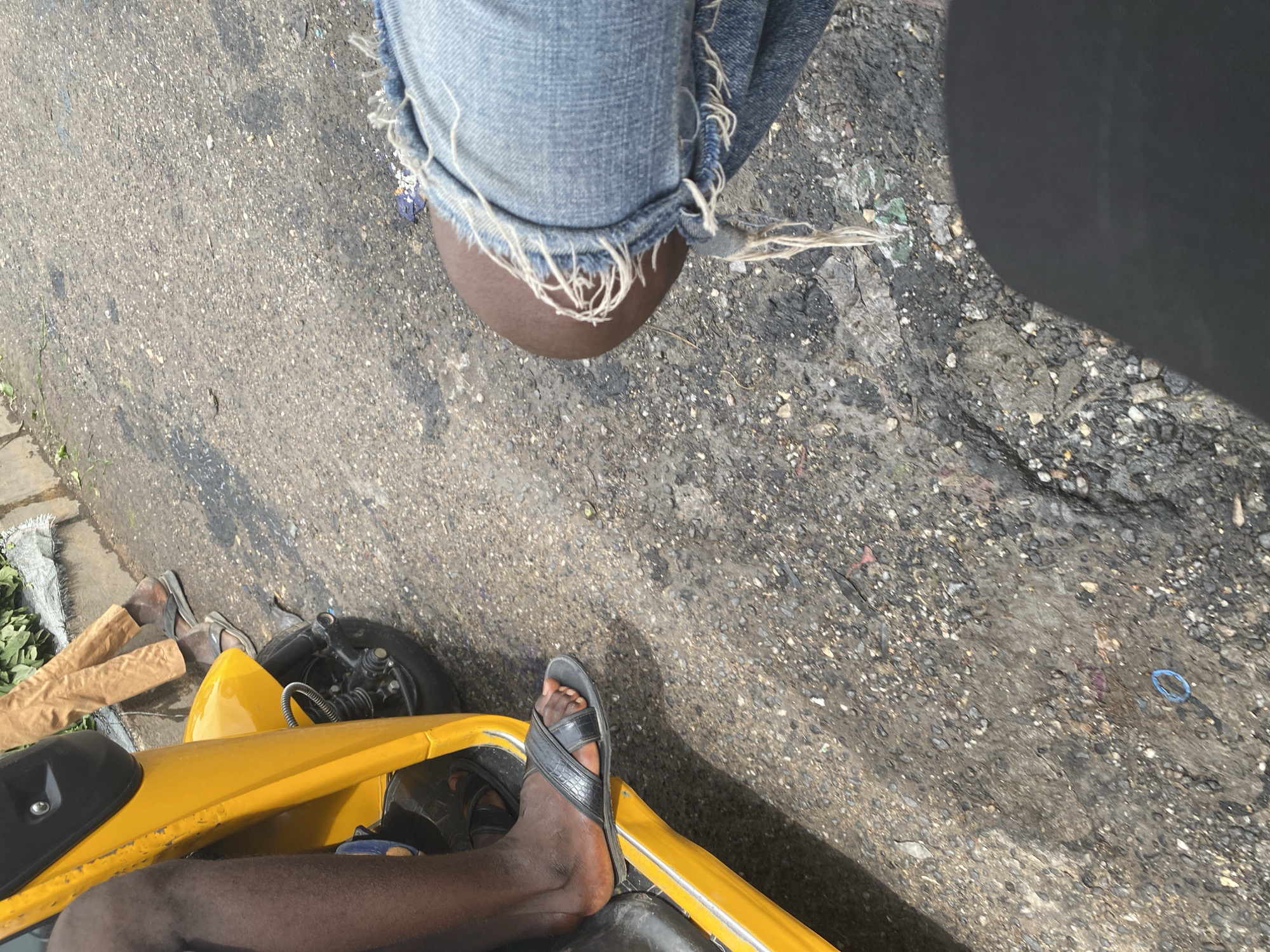 The image shows a ground-level perspective of two people, likely traveling together. One person, wearing distressed denim jeans and dark skin on their lower leg, is positioned higher up in the frame, with their leg resting on an unseen surface. Their jeans appear torn at the hem, with loose threads hanging.
Below this, a yellow vehicle, possibly a tricycle or motorcycle taxi, is partially visible. A person's dark leg, wearing a black sandal with a textured strap, is resting on the yellow body of the vehicle. Another leg, also with a sandal, is visible further to the left, along with two brown paper-wrapped items. Green foliage is visible to the far left of the frame, suggesting an outdoor setting.
The ground beneath is asphalt, with visible gravel and some scattered debris, including a blue rubber band. The lighting suggests it is daytime, with shadows indicating the sun is present, but the overall brightness is moderate. The scene appears to be a street or roadside in Jalingo, Nigeria, given the context. The individuals seem to be in transit, possibly as passengers or operators of the yellow vehicle. There is no visible text in the image.