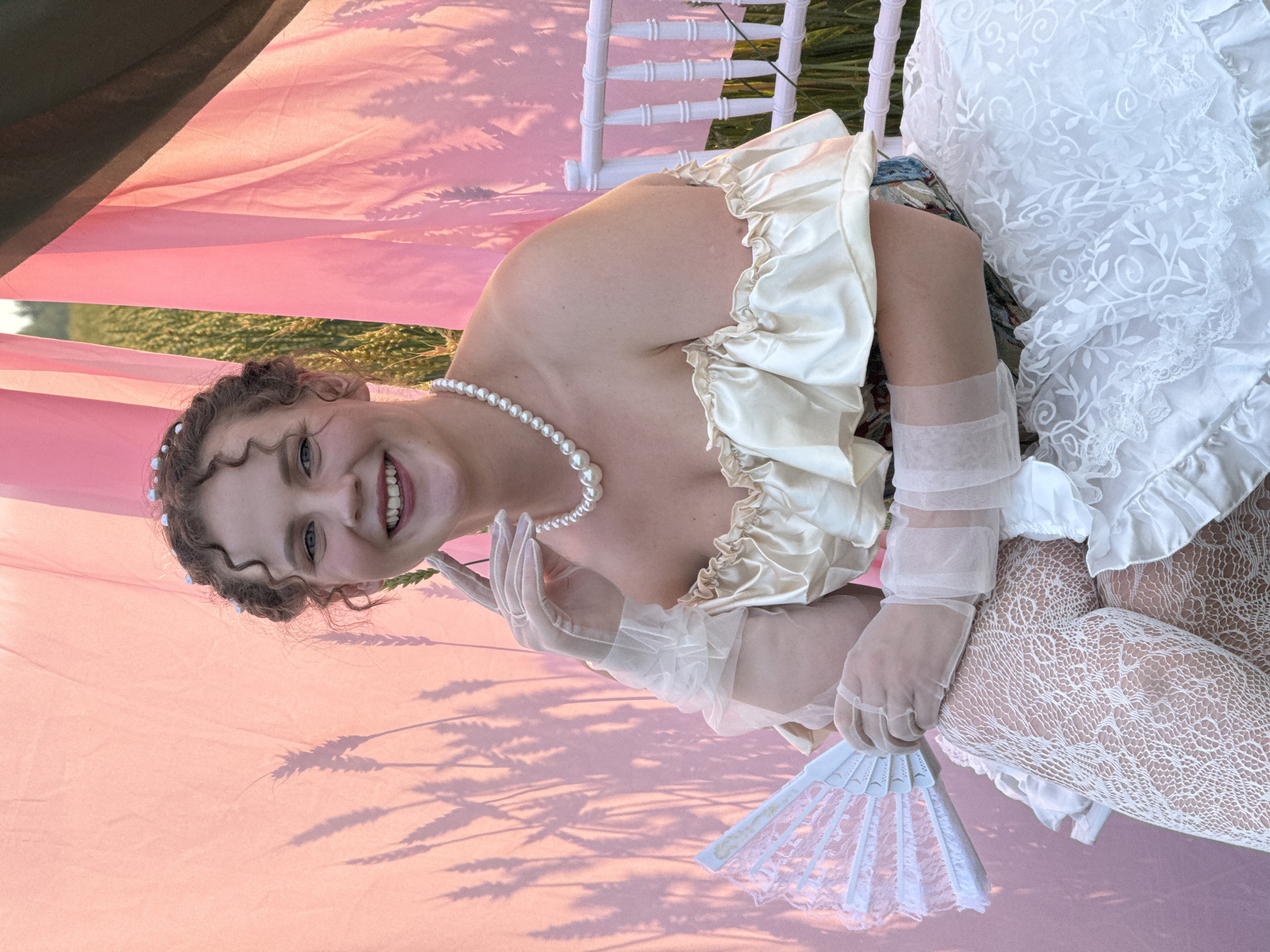 A young woman with curly brown hair is seated on a white chair outdoors. She is wearing an off-the-shoulder cream-colored dress with ruffled details, white lace stockings, and sheer white gloves. A pearl necklace adorns her neck. She holds a white lace fan in her left hand. Behind her, a large pink fabric is draped, casting shadows of wheat stalks. The setting appears to be a field, potentially in or near Malyi Bukor, Russia, given the presence of wheat and the outdoor environment. The lighting suggests it is late afternoon or early evening.
