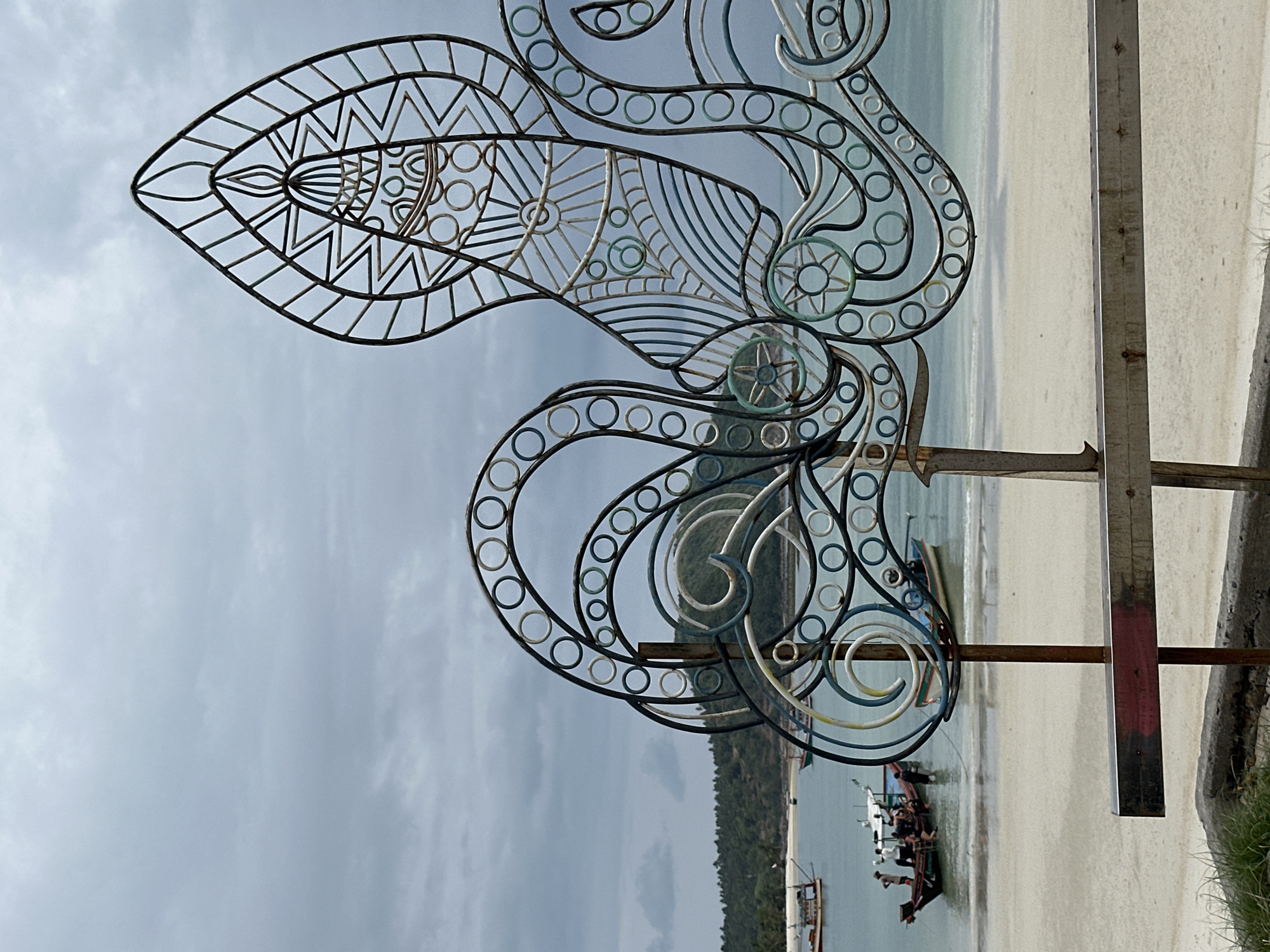 The image displays a large, ornate metal sculpture set against a coastal landscape under an overcast sky. The sculpture, likely made of wrought iron, occupies the foreground and features intricate patterns including an elongated, eye-like shape at the top-left, filled with geometric lines and zigzags. Below this, the metalwork consists of flowing curves, spirals, and numerous small, concentric circles. Some sections of the sculpture exhibit a light blue or greenish patina, possibly from oxidation or a painted finish. On the far right, a wooden or metal post, part of the sculpture's support structure, is visible.
In the background, a sandy beach stretches along the shore, meeting calm, light blue water. Several longtail boats, typical of Thailand, are dotted in the water close to the beach, with some individuals visible on or near them, suggesting activity related to local transport or fishing. Further in the distance, a lush, green, forested coastline rises. The sky is covered with soft, grey clouds, indicating an overcast daytime. The scene combines artistic craftsmanship with the natural beauty and local maritime life of Ko Pha-ngan, Thailand. No text is visible in the image.