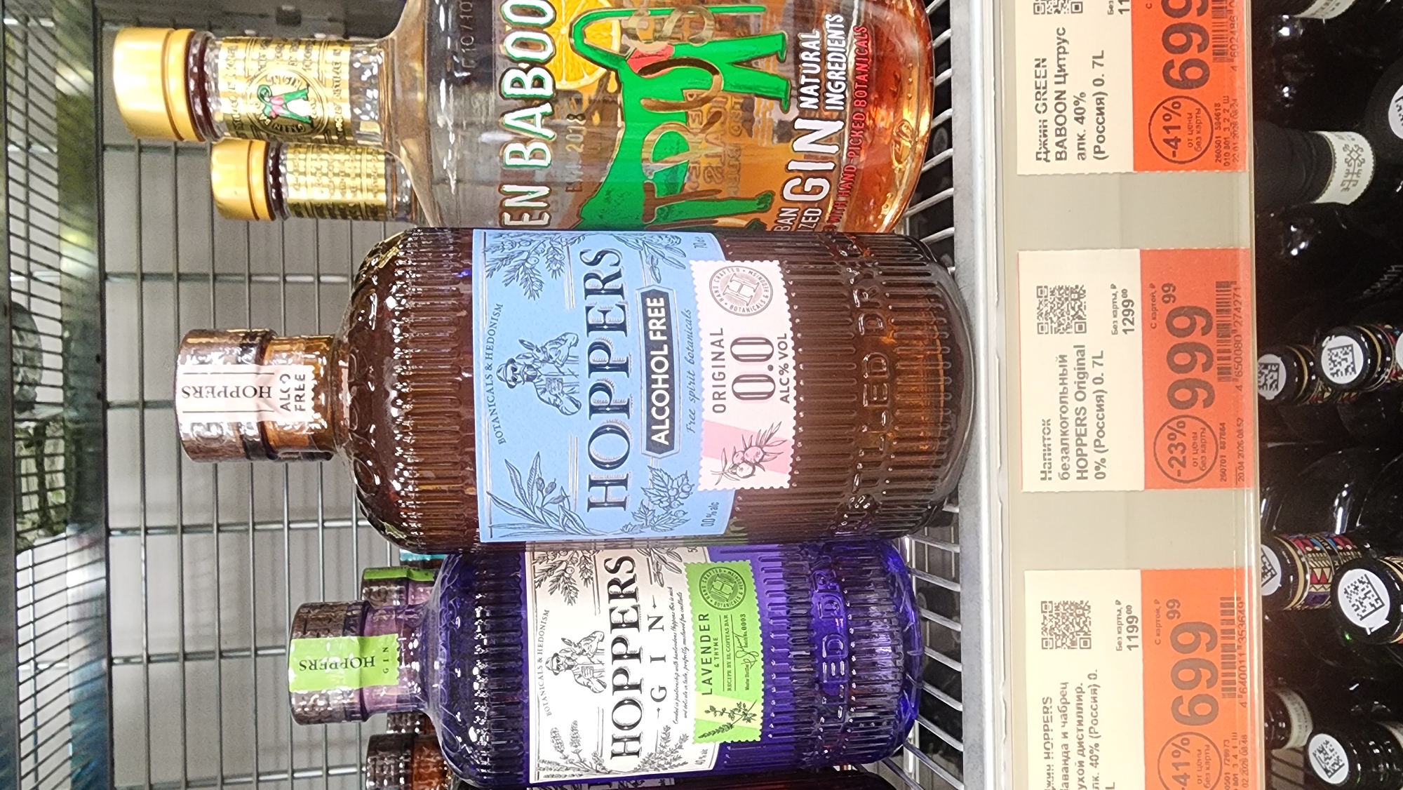 The image shows a shelf in a store, possibly a supermarket, located in Moscow, Russia, displaying several bottles of gin and non-alcoholic gin alternatives. The primary products are: "GREEN BABOON" gin, "HOPPERS Original 0.0%" alcohol-free gin, and "HOPPERS Gin Lavender & Thyme." Price tags, including promotional pricing, are visible adjacent to the bottles. The image captures a typical retail environment where alcoholic and non-alcoholic beverages are sold.