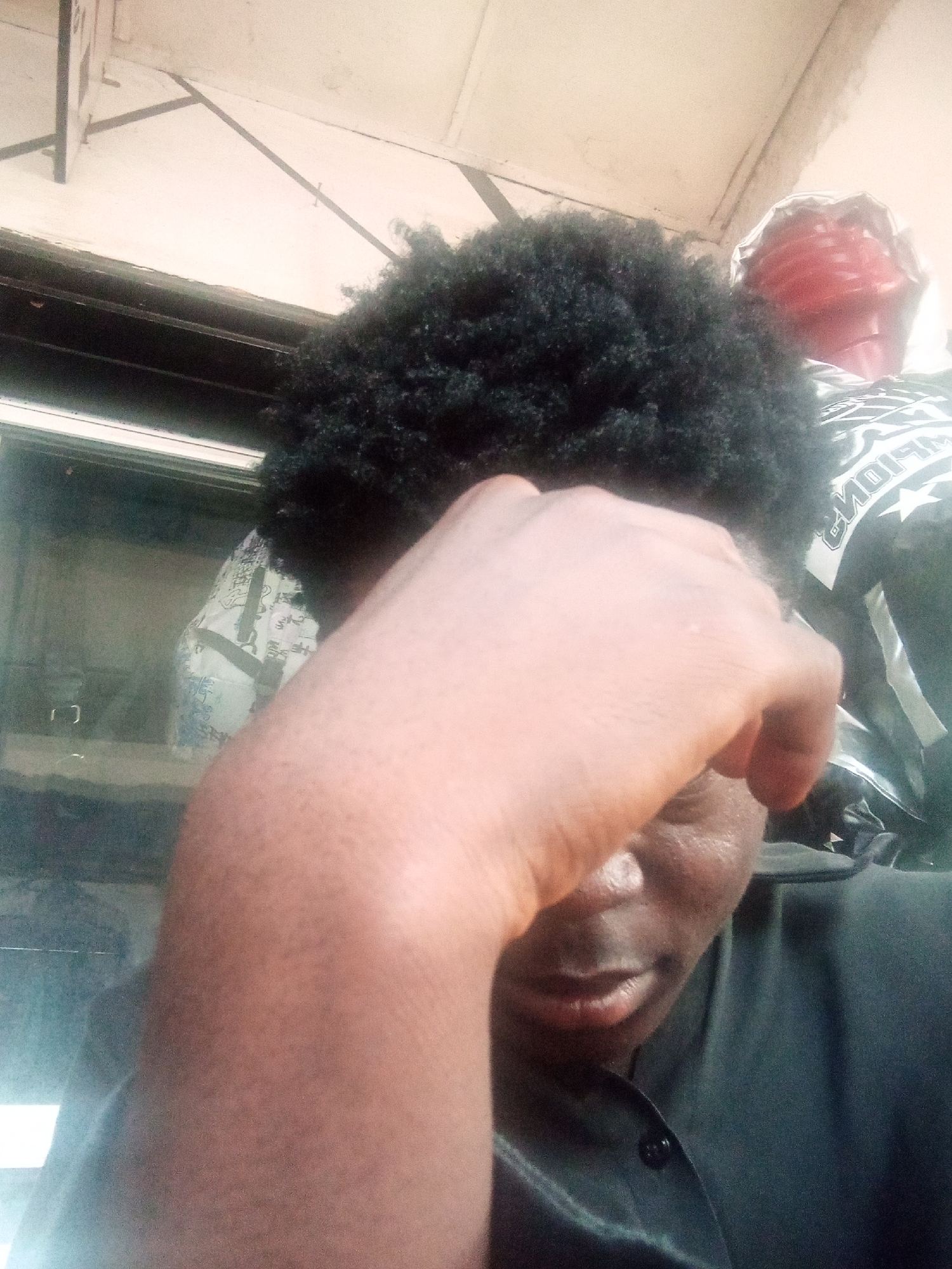 A person is captured in a close-up, low-angle shot, holding their hand to their forehead. Their dark, curly hair is prominent, and they are wearing a dark collared shirt. In the background, a red, foldable object is visible, along with a black and white patterned garment. The environment appears to be indoors, with a light-colored ceiling and some visible structural elements. The lighting is bright, casting a slight glare on the scene. The image quality is somewhat grainy, suggesting it may have been taken with a mobile device in less than ideal lighting conditions. The overall impression is one of introspection or perhaps discomfort. The location is identified as Bukuru, Nigeria.