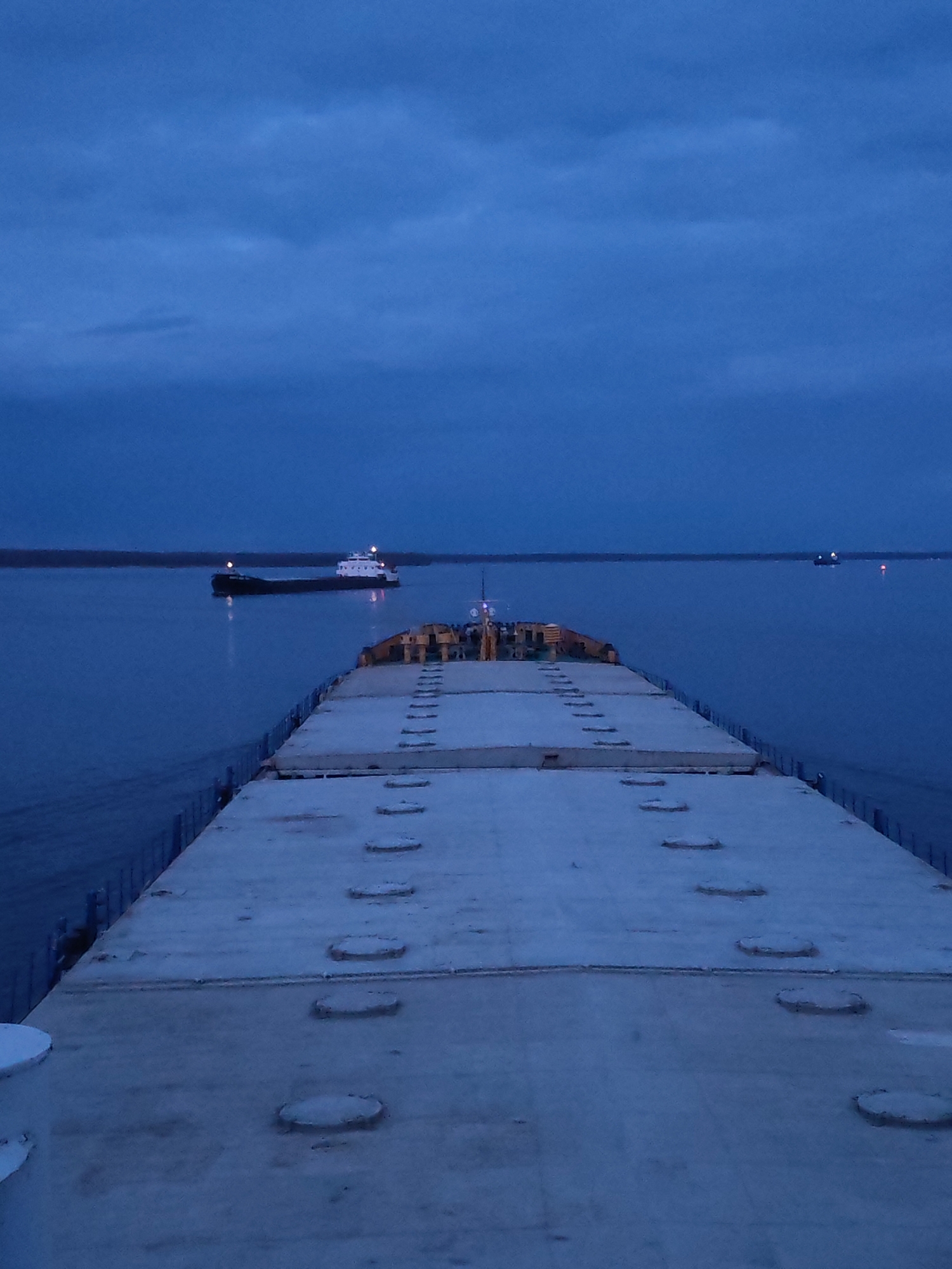 The image presents a wide, low-light view from the stern of a large vessel, likely a barge or cargo ship, on a substantial body of water near Gorushka, Russia. The foreground shows the broad, flat deck of the viewer's vessel, covered with evenly spaced, circular hatch covers running down its length. The deck surface appears light-colored, possibly due to low light or a thin layer of frost.
In the mid-ground, to the left, another large cargo ship is visible, either navigating or anchored. It has a dark hull and a lighter-colored superstructure, with several illuminated lights on its deck and mast reflecting on the calm, dark blue water. Further in the distance, a faint light source is visible on the water to the right, suggesting another distant vessel or buoy.
The water itself is a deep, tranquil blue, mirroring the color of the overcast sky above. A dark, indistinct landmass or shoreline stretches across the horizon. The sky is a uniform, dark blue, indicating either dusk or dawn, often referred to as the "blue hour," with subtle cloud formations adding texture. The artificial lights on the vessels stand out against the dim natural light. No text is visible in the image. The scene evokes a quiet, solitary atmosphere on a waterway during twilight hours.
