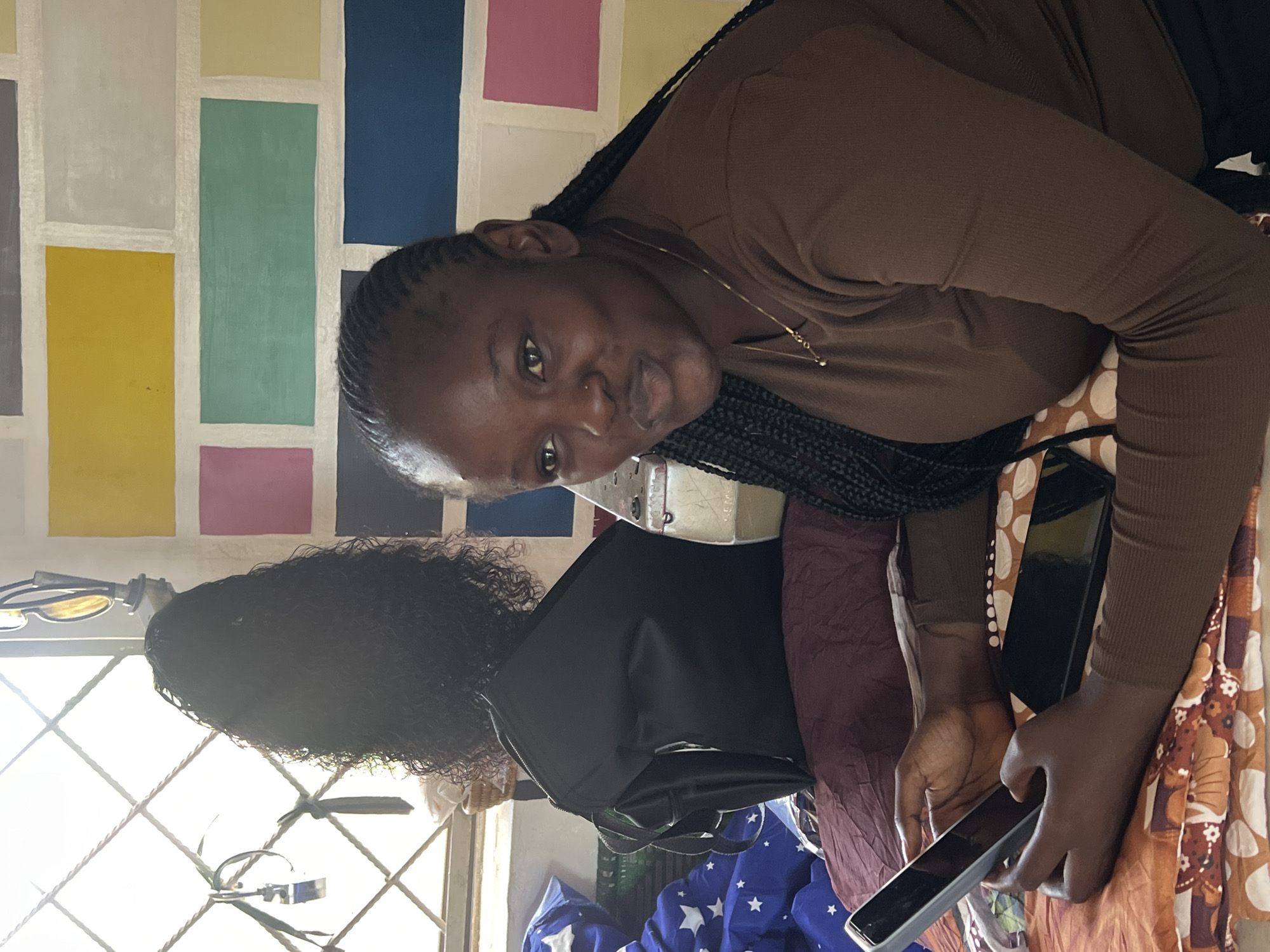 In Jos, Nigeria, a young Black woman with braided hair is seated on a bed, holding a smartphone in her hands. She wears a brown long-sleeved shirt and a delicate gold necklace. To her left, a black backpack is placed on a burgundy pillow, with a black wig displayed behind it. The room is decorated with a vibrantly colored brick-patterned wall. Sunlight streams through a window with a diamond-patterned grate. A blue patterned duvet is visible on the bed.