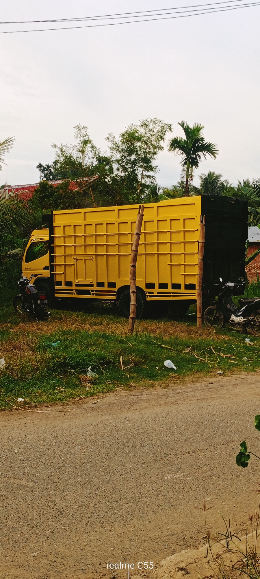 A side view of a bright yellow truck with a black cargo box is parked on a grassy shoulder next to a dirt road. The truck's bed has a grid-like structure. A dark-colored motorcycle is parked to the left of the truck, and another motorcycle is parked to the right. Two wooden poles stand vertically in front of the truck's cargo box. The background consists of lush green foliage, including palm trees and bamboo, with a glimpse of a red-tiled roof in the upper left. The sky is overcast. The image appears to have been taken in a rural area, likely in the city of Lhokseumawe, Indonesia. The lower right corner of the image contains a watermark "realme C55".