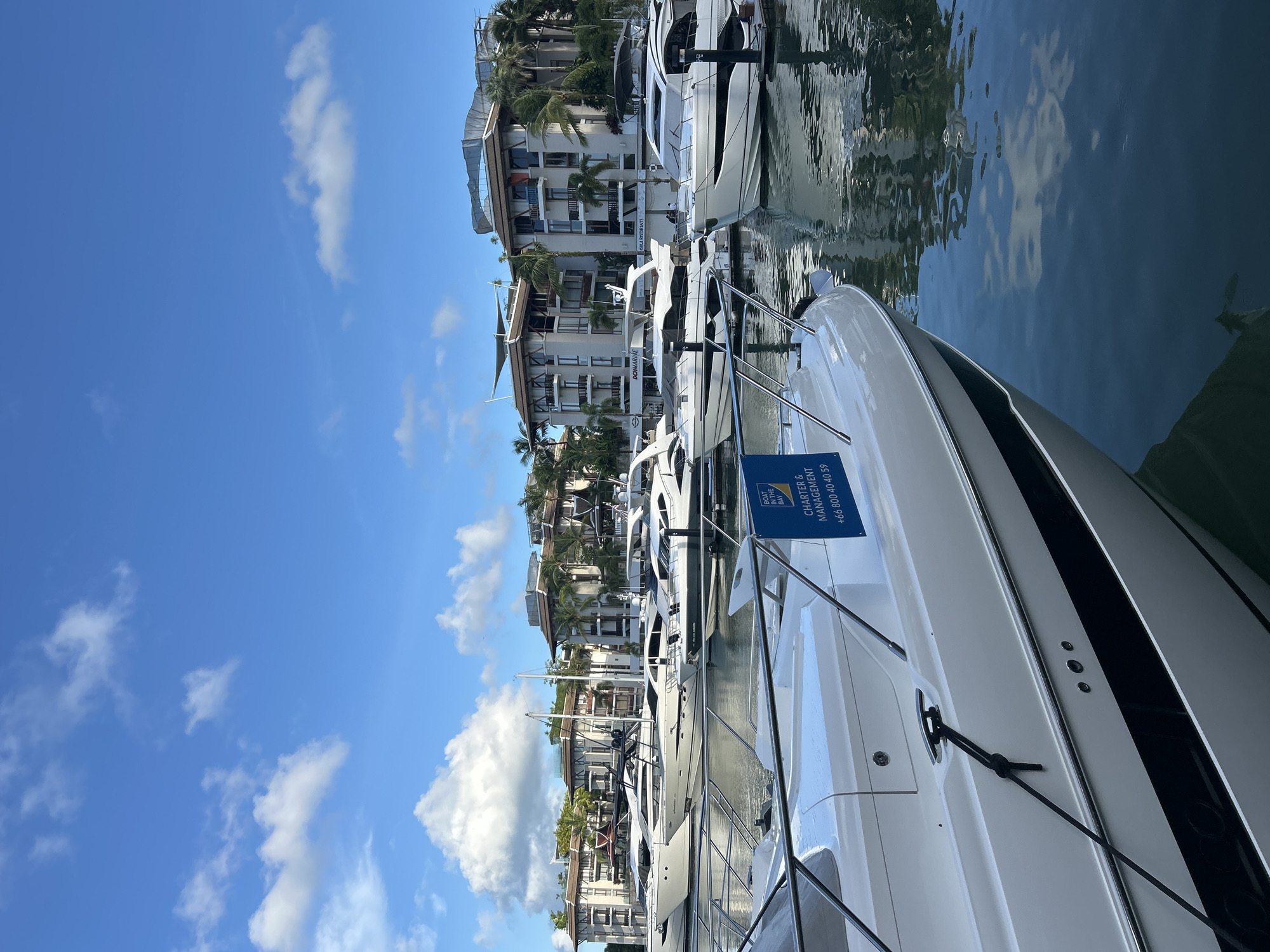 An outdoor, medium-shot, eye-level, full-color photograph depicts a marina with several luxury yachts docked in the water. The scene is located in the city of Ko Kaeo, Thailand. In the foreground, the bow of a white speedboat is prominently featured, with a blue sign attached to its railing that reads "BOAT IN THE BAY CHARTER & MANAGEMENT +66 800 40 40 59". The water is calm, reflecting the blue sky, white clouds, and the surrounding buildings and yachts.
In the mid-ground, a row of sleek, modern yachts are moored side-by-side. These yachts are predominantly white with dark accents. Behind the yachts, a line of multi-story buildings with beige facades, dark roofs, and balconies can be seen. Palm trees are interspersed among the buildings, suggesting a tropical or subtropical climate. The sky is a vibrant blue, dotted with fluffy white clouds. The lighting suggests it is daytime, likely morning or afternoon, with the sun casting soft shadows. There are no people visible in the frame.
