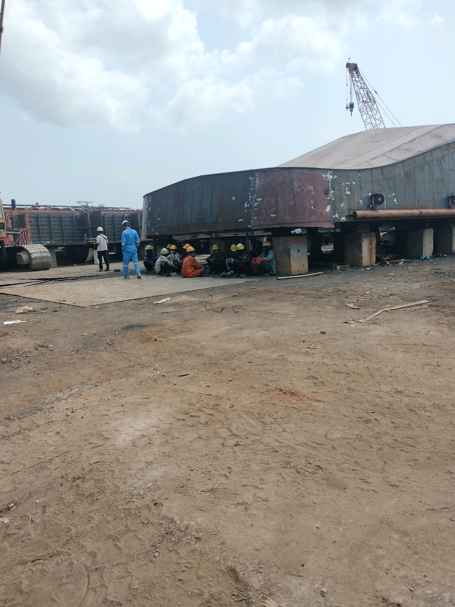 The image depicts an industrial scene, likely within a shipyard or heavy construction area in Batam, Indonesia. In the foreground, a dusty, unpaved ground surface is visible, marked with tire tracks and footprints. A large, curved metallic structure, possibly a section of a ship or industrial tank, is elevated on concrete blocks in the middle ground.
Several individuals are present. Two men are standing near the metallic structure, one in a blue uniform and white hard hat, and another in lighter clothing and a white hard hat. A group of about eight people, all wearing hard hats, are seated on the ground in front of the elevated structure. Some are wearing orange or green work clothing, while others are in dark attire. They appear to be taking a break or conversing.
In the background, a large crane with a long boom is visible against a cloudy sky. To the left, the tracked portion of a heavy construction vehicle is partially visible. Additional structures, possibly more industrial equipment or parts of a facility, are also present in the distant background. The overall environment suggests a working shipyard with ongoing construction or repair activities.