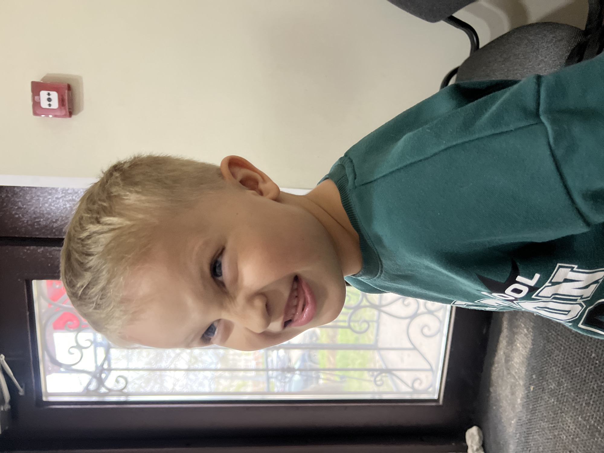 A young boy with blond hair and blue eyes is smiling and looking downward. He is wearing a green sweatshirt with white lettering. He is standing near a window with a dark frame and decorative metal bars. Outside the window, there is greenery. A red fire alarm box is mounted on the wall. Part of a gray chair is visible in the background. The location appears to be indoors. The city and state are unknown.