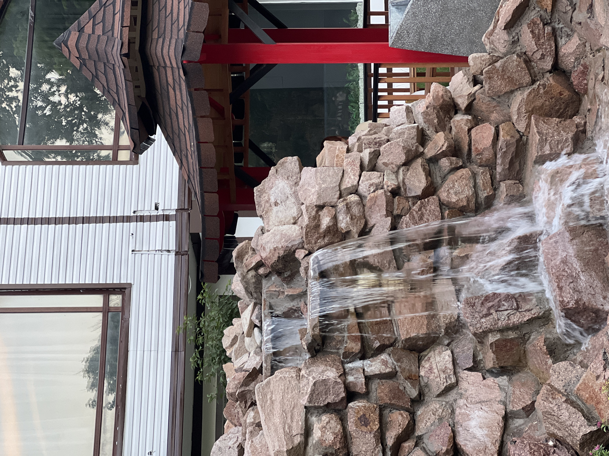 The image depicts an outdoor scene featuring a multi-tiered rock waterfall. Water cascades over large, rough-hewn stones that are predominantly reddish-brown in color, creating a naturalistic effect. Behind the waterfall, a building with white siding and a brown-trimmed, angled window is visible. To the right of the waterfall, a structure with a pagoda-style roof, tiled in shades of brown and red, is supported by bright red posts. The background also shows some greenery and a wooden fence. The scene appears to be located in Shymkent, Kazakhstan, possibly in a garden or park setting. No people are clearly visible in the image.
