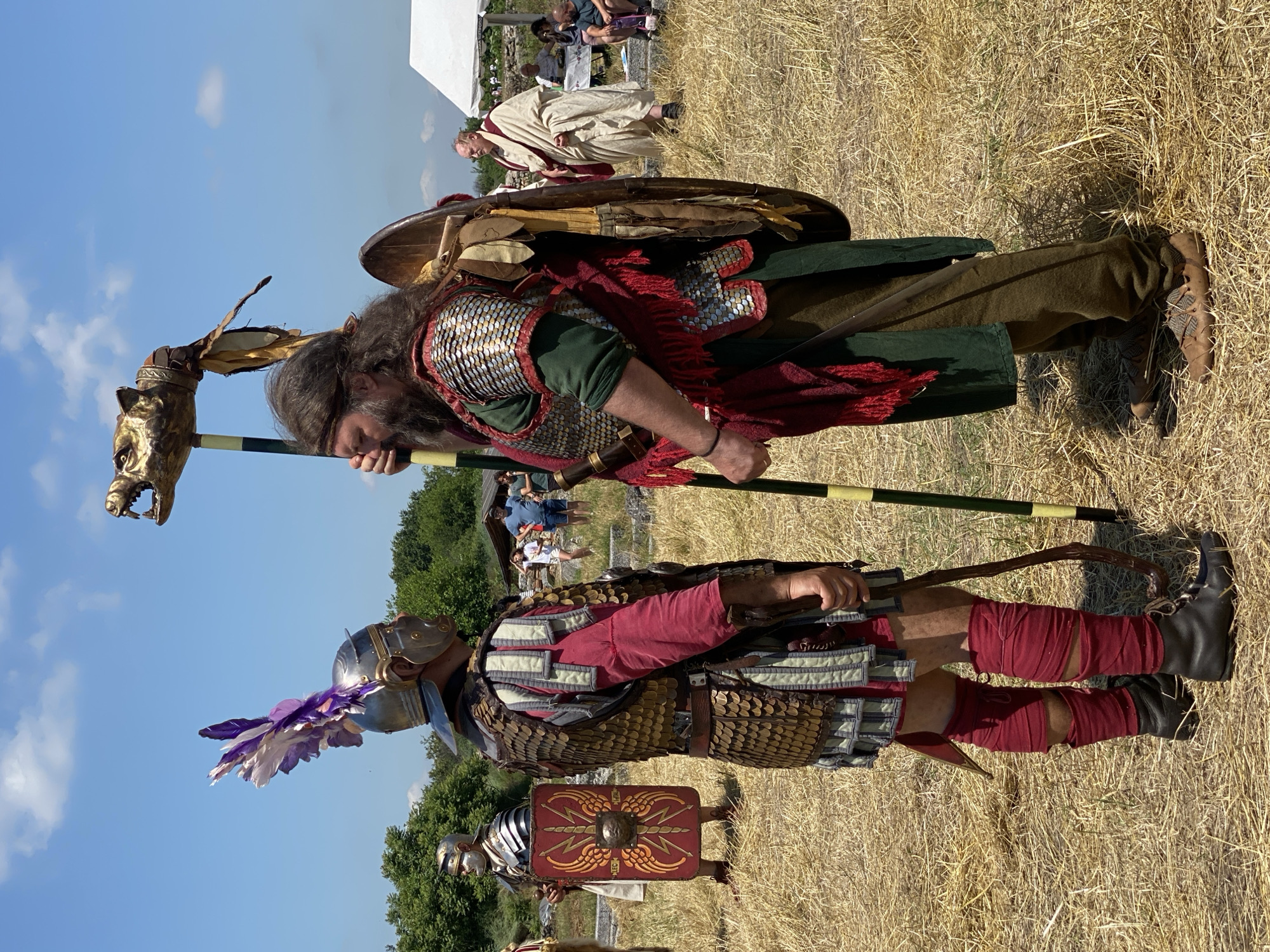 This image captures a historical reenactment scene outdoors in Gigen, Bulgaria, under a clear blue sky with sparse white clouds. The ground is covered in dry, golden straw or hay.
Two primary figures are positioned centrally, facing each other in apparent interaction.
On the left, a man is dressed as a Roman soldier. He wears a metallic galea-style helmet adorned with a large, upright plume of purple and white feathers. His body is covered by bronze-colored scale armor (lorica squamata) worn over a red long-sleeved tunic. Light grey and blue pteryges (protective strips) are visible at his shoulders and waist. His lower legs are wrapped in red fabric (fasciae), ending in black, low-cut boots. He holds a short wooden staff in his right hand.
On the right, a man with long, dark brown hair and a full beard is dressed as a barbarian or standard bearer. He wears metallic, silver-colored scale armor over a green tunic. A red fringed scarf or cloak is draped over his left shoulder. He holds a tall standard (signum) in his left hand, which features a golden animal head (resembling a wolf or draco) mounted on a pole striped with green and yellow bands. A round, wooden shield, decorated with light-colored leather strips, is secured to his back. He wears olive green trousers and light brown, low-cut shoes with a patterned design.
In the mid-ground to the left, another man in Roman military attire stands, wearing a helmet and scale or mail armor, with his face painted white. He carries a rectangular scutum (shield) with a red background, featuring a golden winged eagle emblem and the letters "LEG" and "MAL."
In the mid-ground to the right, an older man in a white toga with red trim walks away from the central figures towards a white tent. Further in the background, several individuals in modern casual clothing are visible, some near the white tent, others observing the scene. Green foliage and trees form the horizon on the left side of the image.