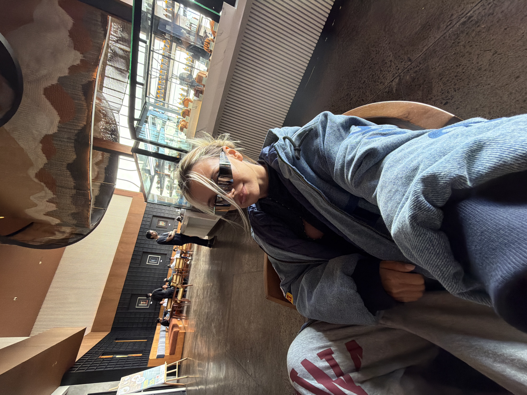 In a spacious, modern indoor cafe, a young blonde woman with striking futuristic sunglasses poses for a selfie. She is seated, wearing a denim jacket over a dark top and grey sweatpants with a red graphic design. The cafe has a high ceiling with an artistic, abstract ceiling installation and a patterned wall. To the right, a glass display case showcases what appear to be pastries or desserts. In the background, several patrons are seated at tables and a man in a black shirt stands near a counter. The floor is made of polished concrete, and warm lighting illuminates the space. The overall ambiance suggests a trendy and contemporary establishment.