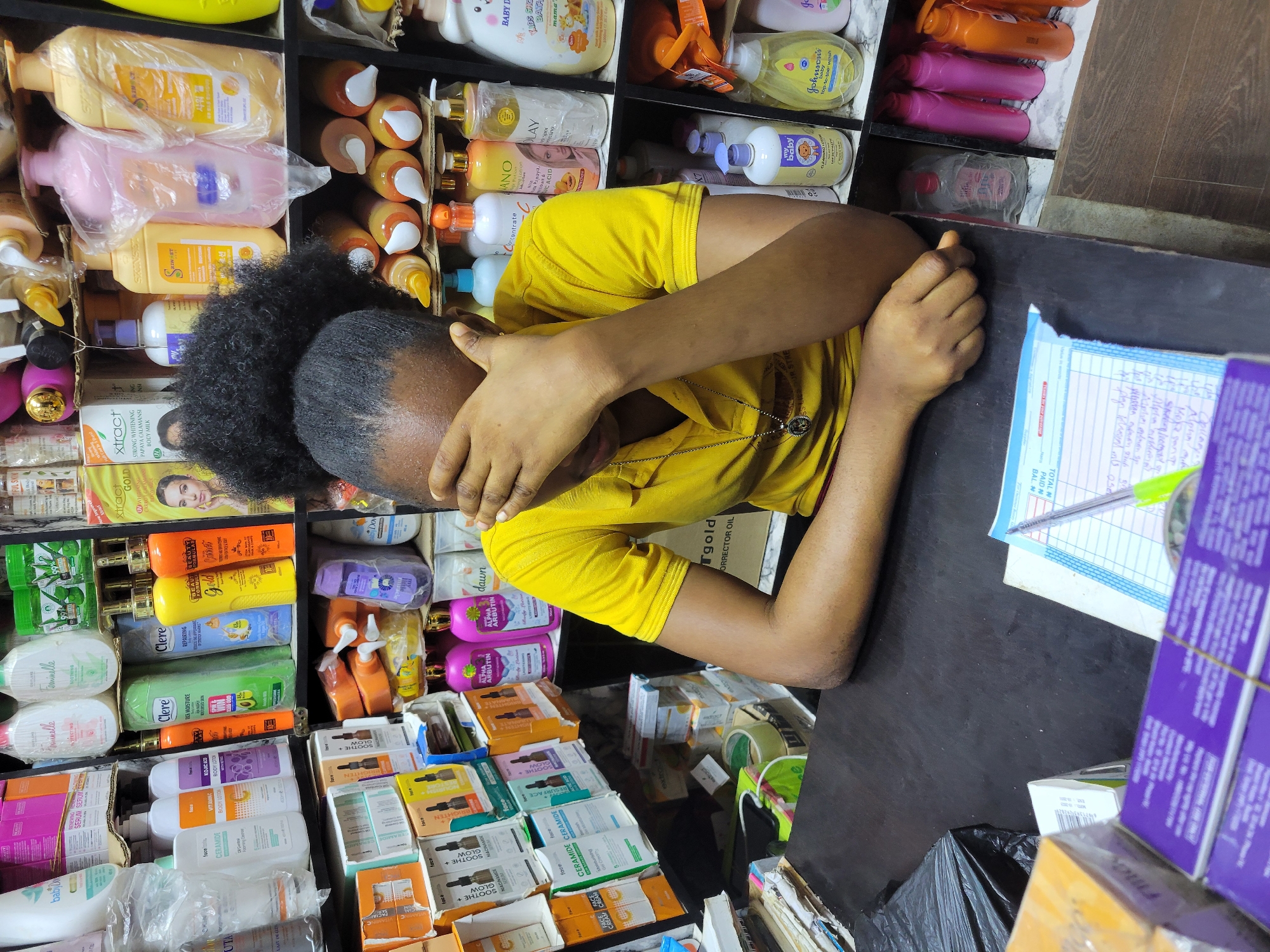 A woman with natural black hair, wearing a yellow t-shirt and a necklace, sits at a counter in what appears to be a small shop in Onitsha, Nigeria. She has her head in her hands, seemingly resting or possibly upset. The counter is cluttered with beauty products, including lotions, creams, and serums from brands like Clere, Feminnelle, and Extract. The shop's shelves behind her are stocked with similar products. There is a pen and a notepad on the counter, along with several boxes stacked on top of each other. A black plastic bag is also visible under the counter.
