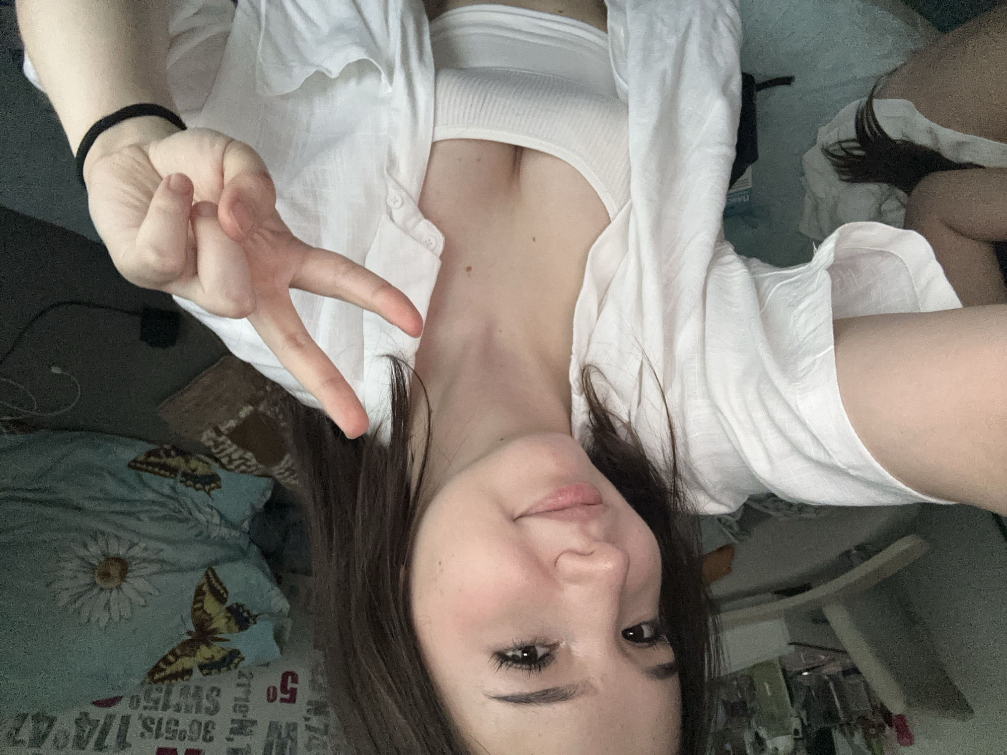 The image shows a young woman taking a selfie indoors, with her upper body filling most of the frame. She is wearing a white tank top under an open white shirt. Her hair is dark brown and falls over her shoulders. She is making a peace sign with her right hand, which is in the foreground. The background is blurred, but appears to be a bedroom with a pillow that has butterflies and a flower pattern. There is also some shelving with items on it. The lighting suggests it is daytime. The woman's expression is a slight smile. The image does not contain any visual information that would allow for the determination of the city of Naberezhnye Chelny, Russia.