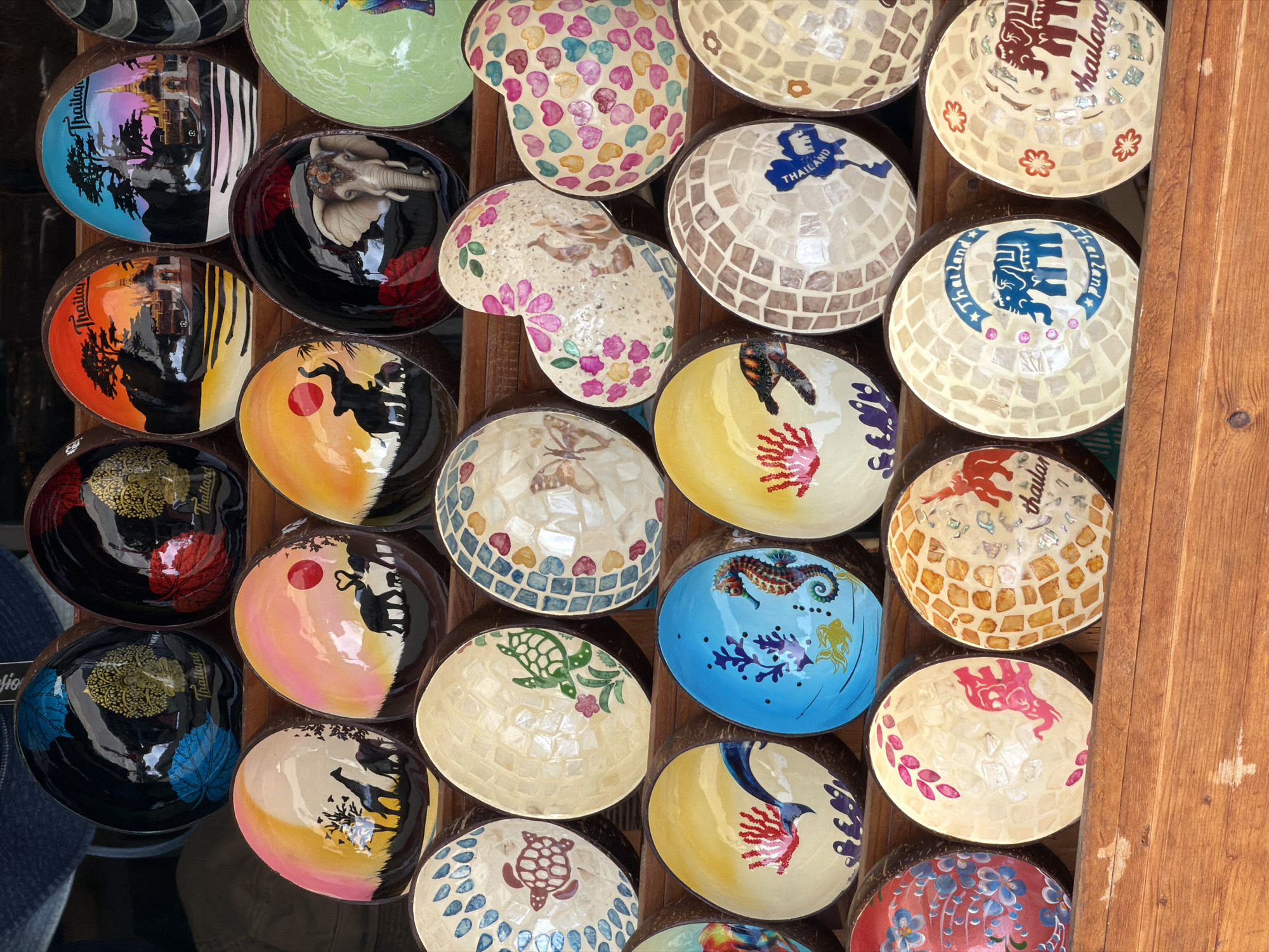 This image depicts a market stall in Baan Plai Laem, Thailand, showcasing a collection of decorative bowls. The bowls are primarily made from coconut shells and are arranged in multiple tiers on wooden shelves. They feature a variety of designs, including painted landscapes with elephants and giraffes, intricate mosaic patterns with shells, and images of marine life like turtles and seahorses. Some bowls are adorned with floral motifs and the word "Thailand." The lighting suggests an outdoor setting, possibly during the day. There are no people visible in the frame.