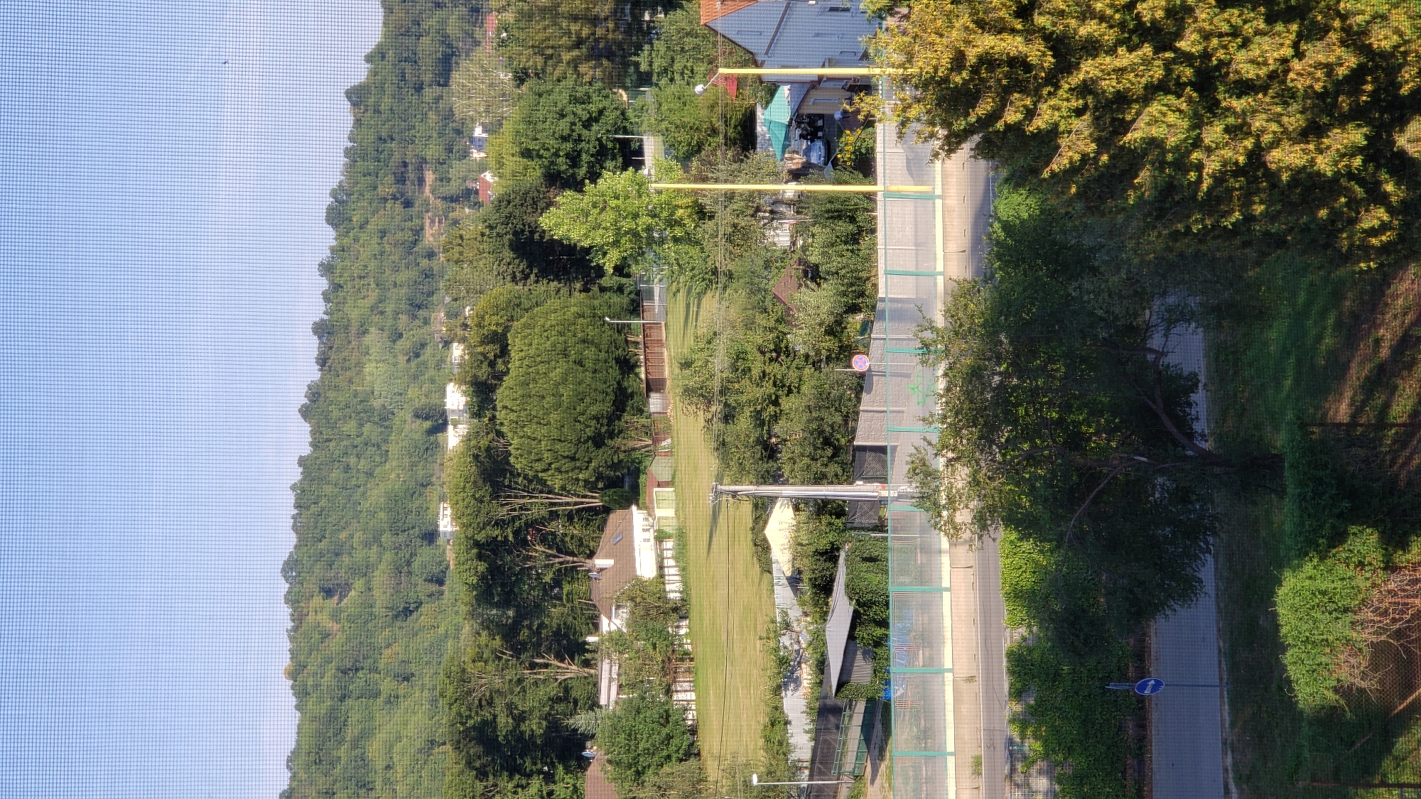 The image depicts a view from a window with a screen, looking out onto a residential area with a tree-covered hill in the background. The scene appears to be located in the city of Prešov, Slovakia, given the context provided.
In the foreground, there are trees and vegetation. A road is visible, with a blue directional sign pointing left. A fence separates the road from a grassy area and more trees. Beyond that, there are several buildings, including houses with white siding and red roofs, and some smaller structures like sheds. A large, well-maintained grassy field is prominent in the middle ground. Further back, the landscape rises into a densely wooded hill, with houses peeking through the trees at the top. The sky is clear and blue. There are no people visible in the image. The overall impression is one of a quiet, suburban setting on a sunny day.