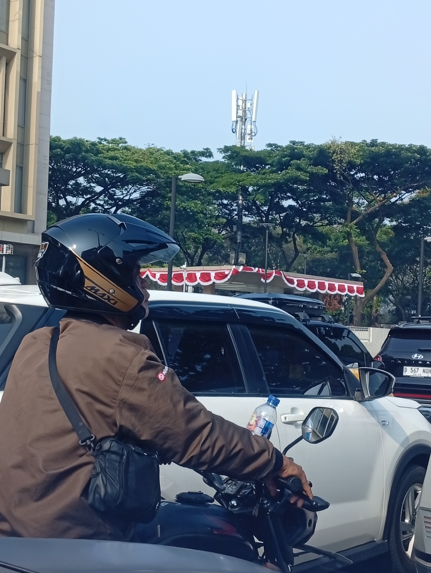 A person wearing a black helmet with gold accents and a brown jacket rides a motorcycle in traffic. The person is looking to the right, with a black bag slung across their chest. A white car is to the right of the motorcycle, and another car is visible behind it. In the background, there are trees, a building, and a cell tower. The scene is set on a street in Lengkong Kulon, Indonesia.