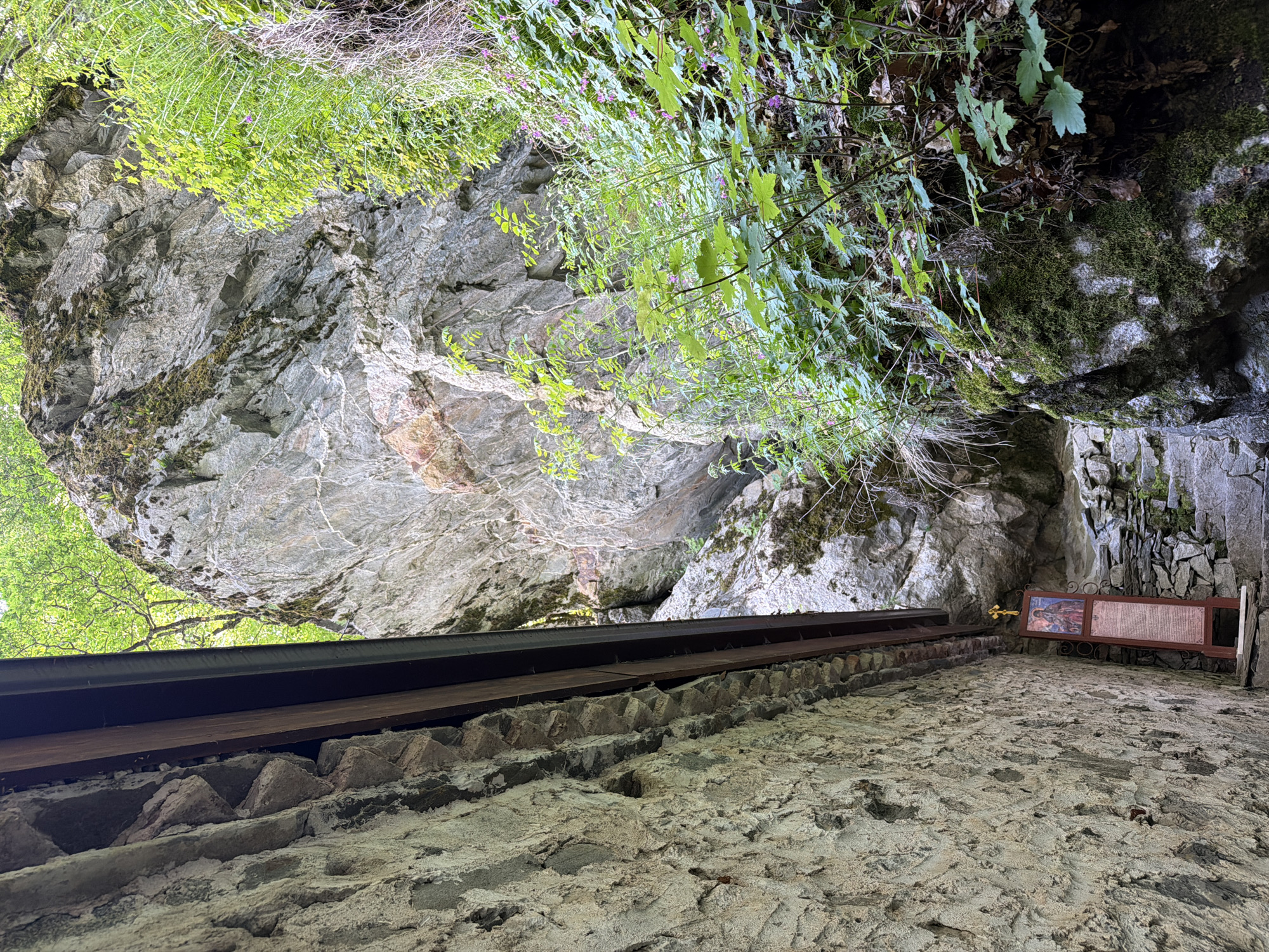 View looking up at a rocky overhang in Govedartsi, Bulgaria. The overhang is part of a natural rock formation, with visible strata and moss growth. Green foliage hangs down from the top of the rock formation. Below the overhang, a small structure with a dark metal roof, wooden beams, and a stone wall. To the right of the structure, an ornate sign with a religious image and text is mounted on the wall. The ground in the foreground is stone-paved.