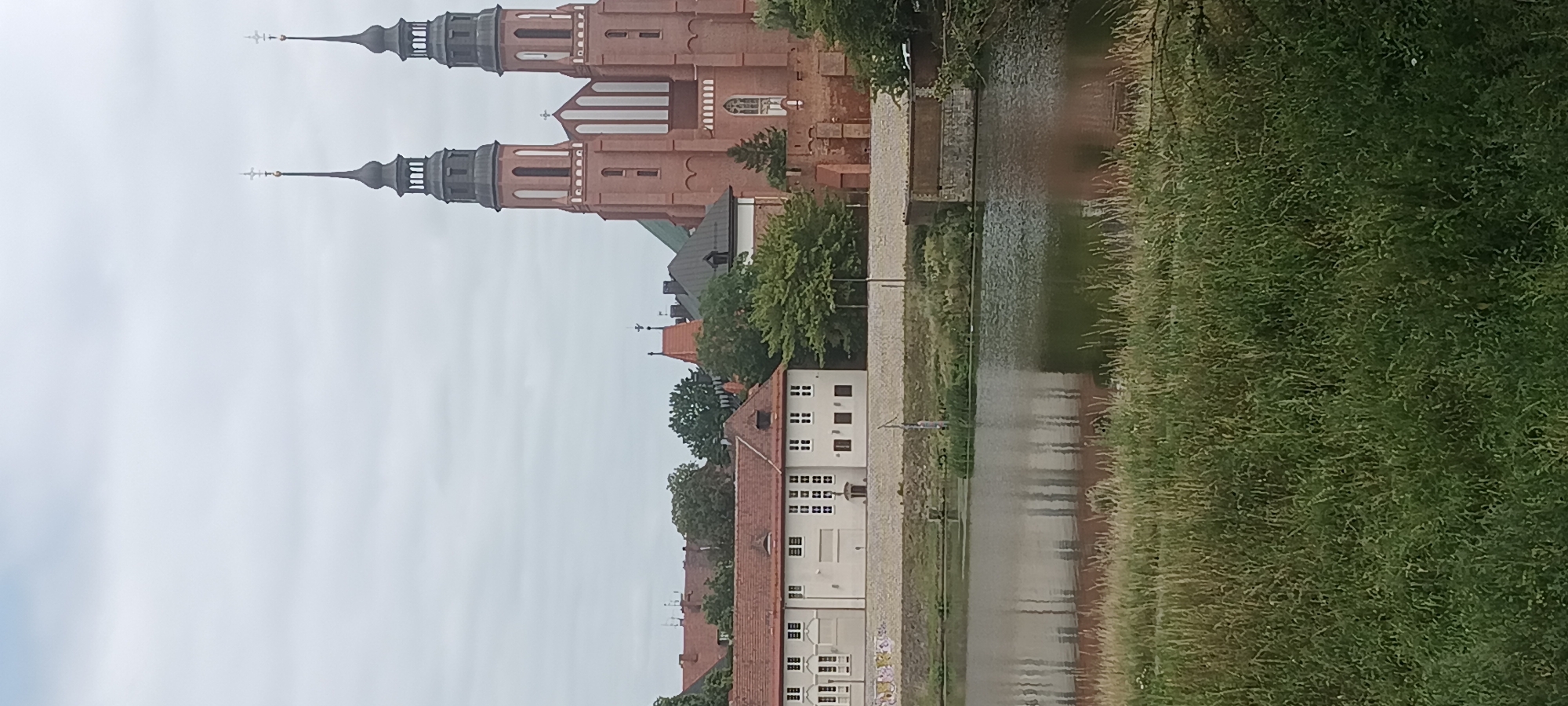 The image depicts a view of the Opole Cathedral in Opole, Poland. The cathedral, a prominent brick building with two tall spires topped with crosses, is situated behind a body of water. In the foreground, tall green grass lines the bank of the water, which reflects the sky and the surrounding buildings. To the left of the cathedral, a long, light-colored building with a red-tiled roof is visible, featuring rows of windows. Trees are interspersed throughout the scene, adding to the natural landscape. The sky is overcast with clouds.