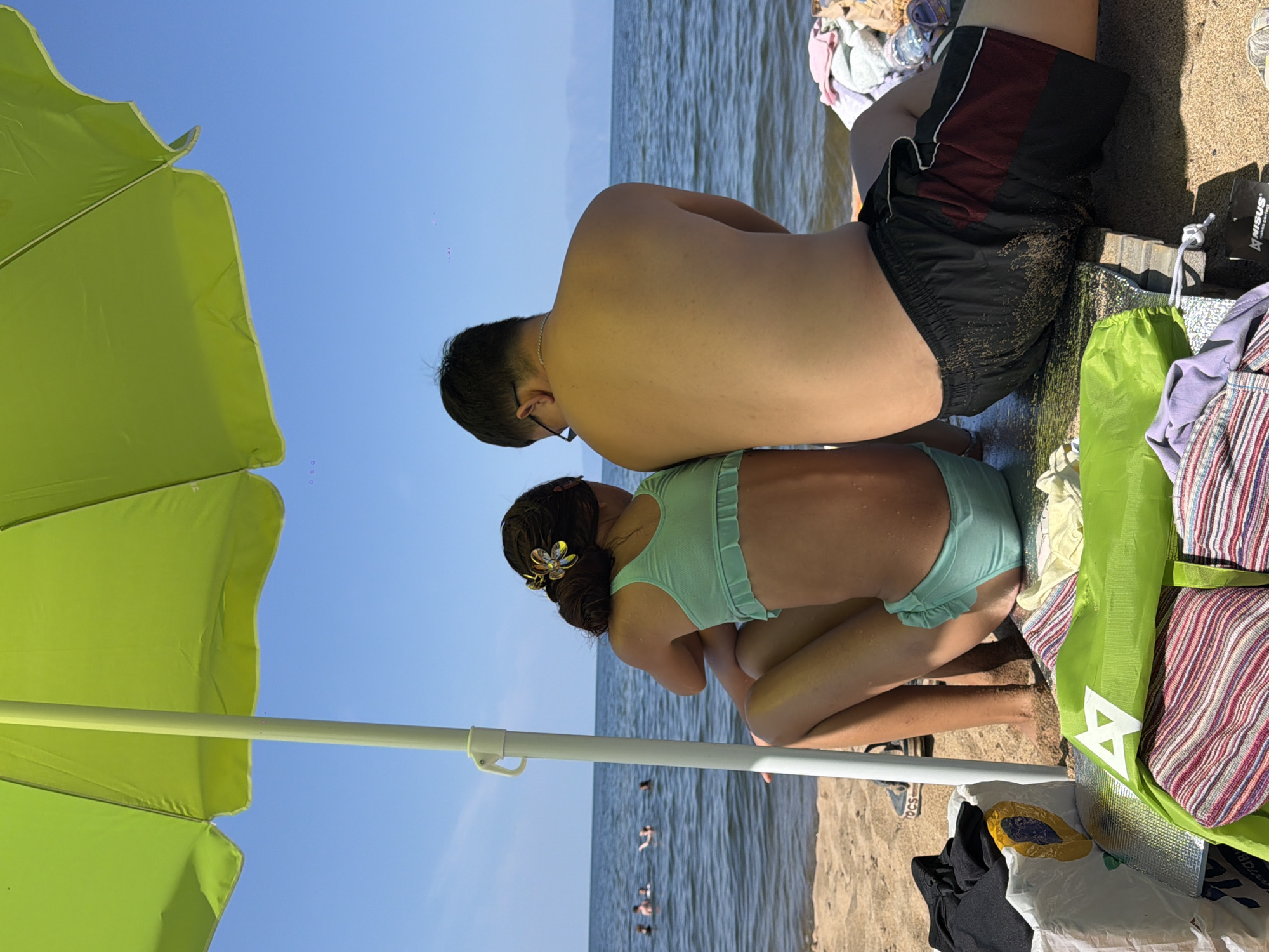 Two people are sitting on a sandy beach next to a body of water. A bright green umbrella is partially visible in the foreground, angled upwards and to the left. The person in the background, viewed from the back, is wearing dark swim trunks with a maroon stripe and appears to be an adult. They are wearing glasses. The person in the foreground, viewed from the side, is a child wearing a light teal bikini. They have a flower in their dark hair, which is pulled back into a bun.
The water is a calm, rippling blue, and several people can be seen swimming in the distance. The sky is a clear, bright blue, suggesting a sunny day. The sand is golden brown, and there are various items scattered around the people, including towels and bags. One bag is bright green and has a white triangular logo. Other items are a striped blanket or towel. A dark object with a yellow oval sticker is on a white surface near the green bag. The overall scene suggests a casual day at the beach. There is no visible text in the image, aside from the logo on the green bag. The time of day appears to be midday or early afternoon due to the bright sunlight. The location is Maksimiha, Russia.