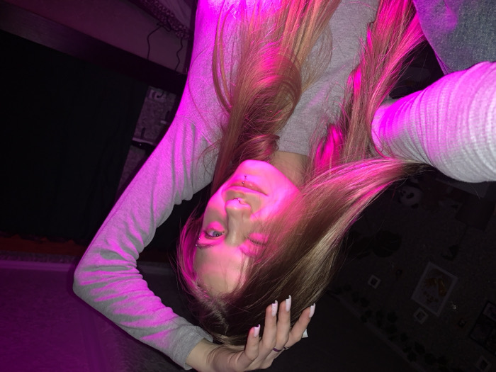 A young female subject with long, straight, blonde hair and fair skin is depicted indoors, purportedly within a residence in the city of Фоки, Russia. She is shown from the chest up, angled slightly, holding a camera to capture a selfie. She wears a light grey, ribbed long-sleeved top. Her left hand is raised, with fingers splayed, resting near the top of her head, displaying manicured nails with white French tips and a dark ring on her left ring finger. Her face is directly illuminated from the left by a strong magenta light source, accentuating her features. She possesses multiple facial piercings, including a septum piercing, a labret piercing below her lower lip, and an eyebrow piercing on her left brow. She is winking with her left eye and has a slight smile. The background is dimly lit, revealing several framed objects on a dark wall and a stuffed panda toy in the lower left corner. Dark furniture is discernible to her right. The scene is predominantly dark, with the magenta light serving as the primary illumination for the subject.